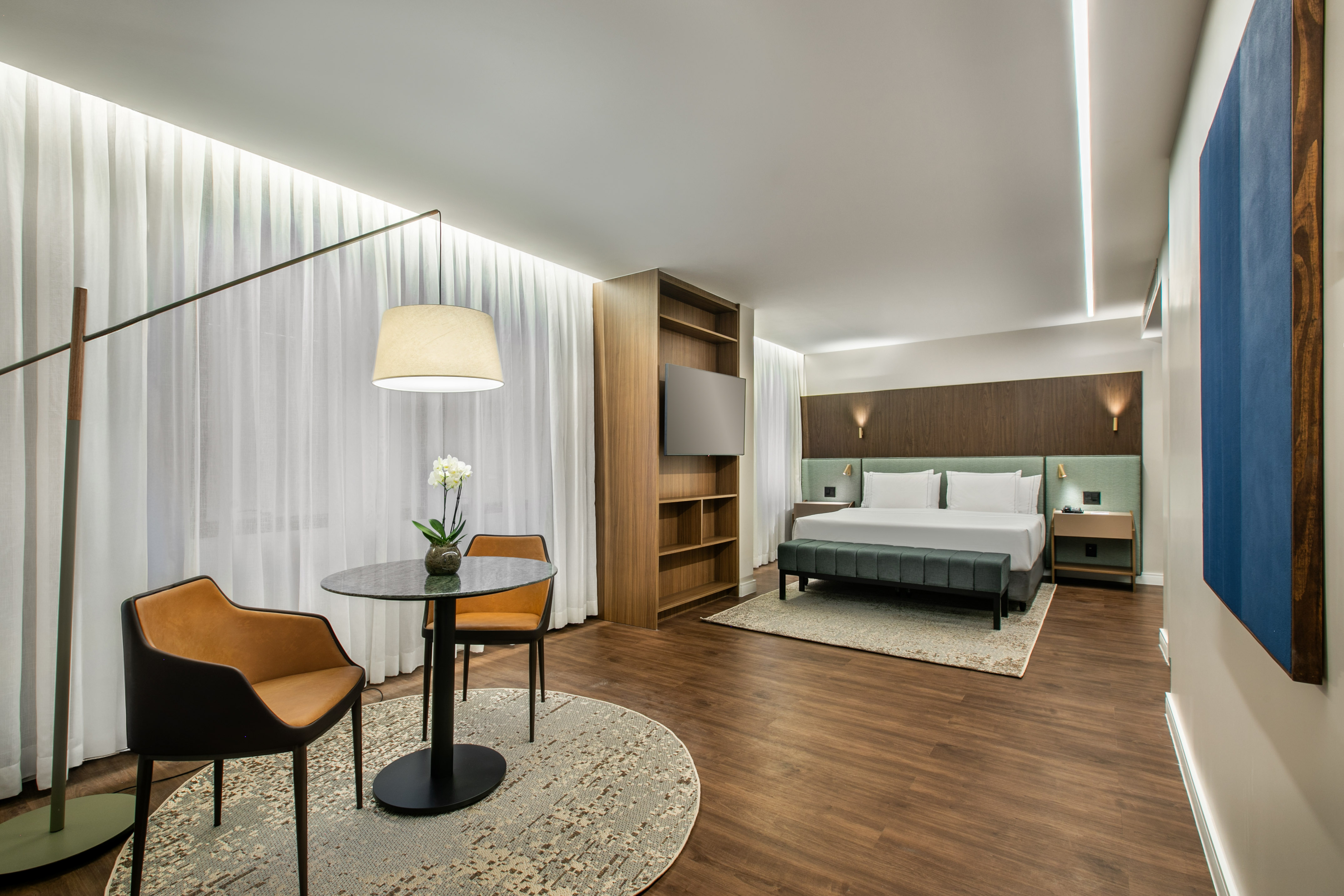 Modern hotel suite with bed, comfortable seating, wooden floors, and sheer curtains.