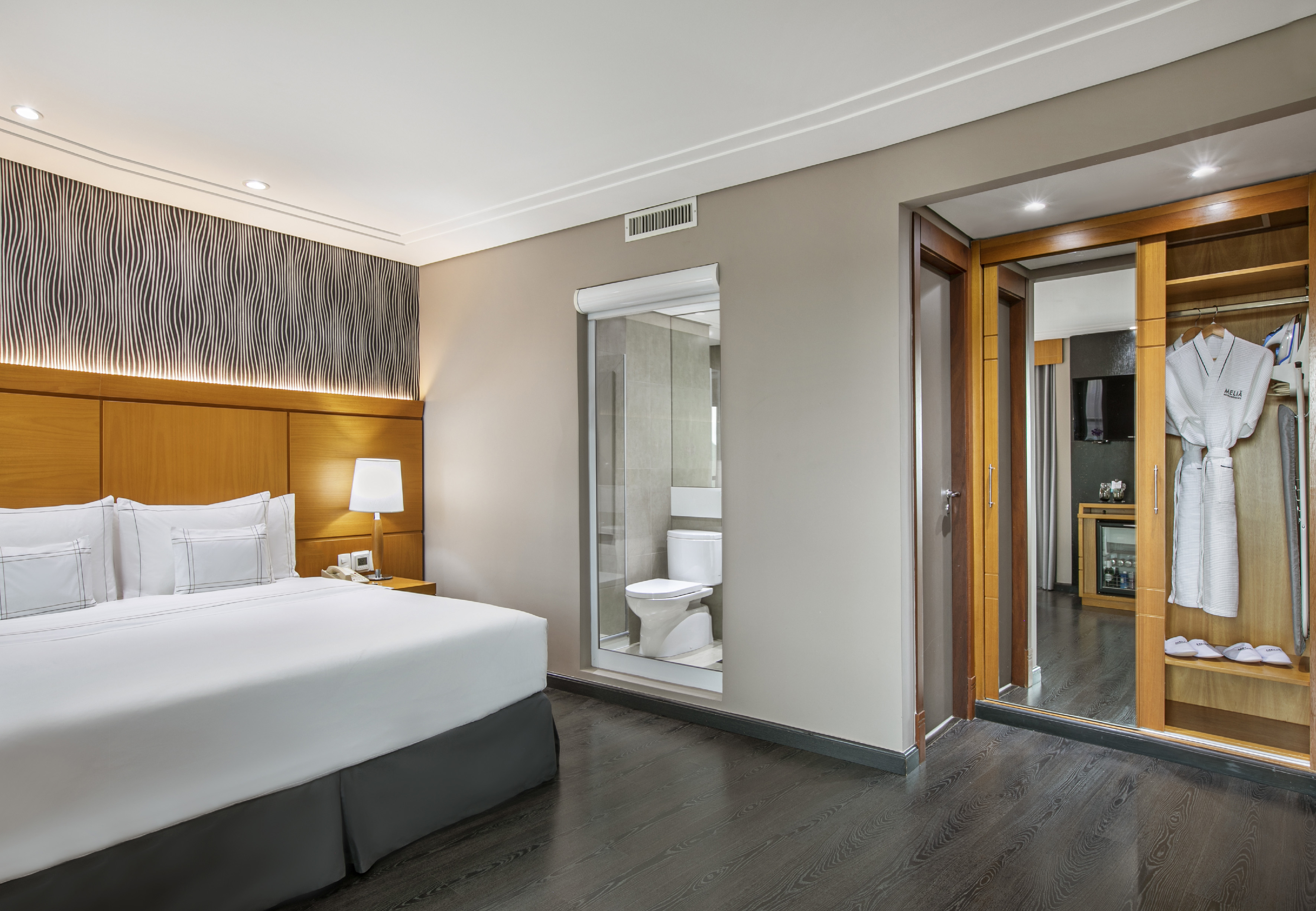 Modern hotel room with king bed, glass-enclosed bathroom, and wardrobe.