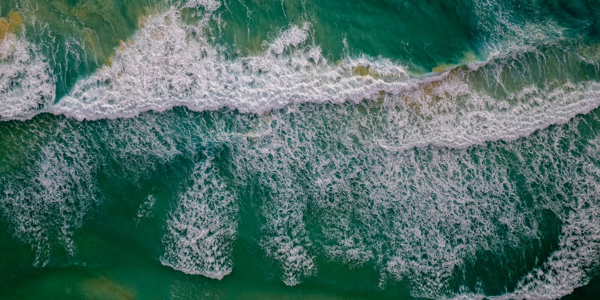 Aerial view of turquoise ocean waves with white foam near sandy beach.