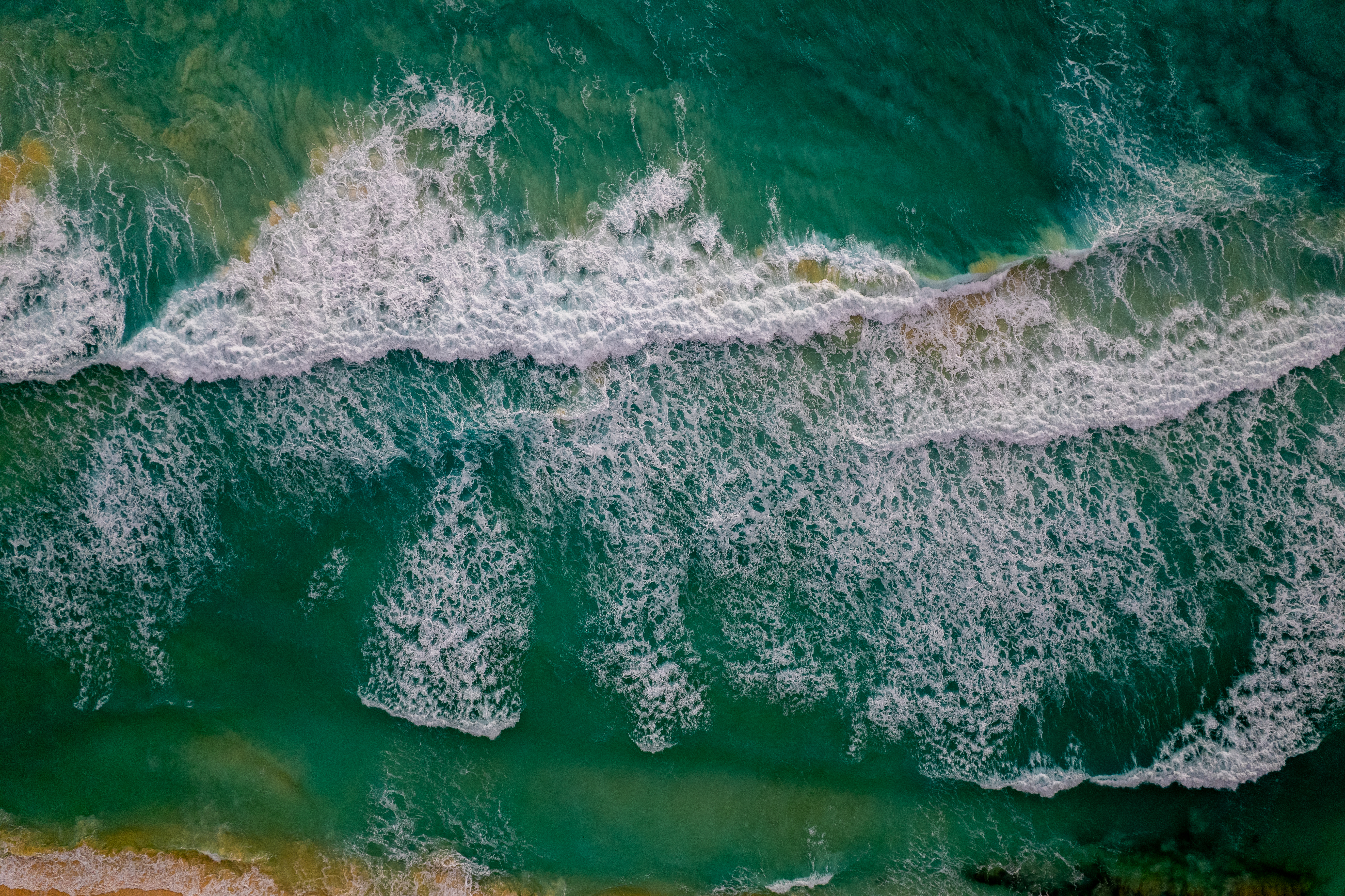 Aerial view of turquoise ocean waves with white foam near sandy beach.
