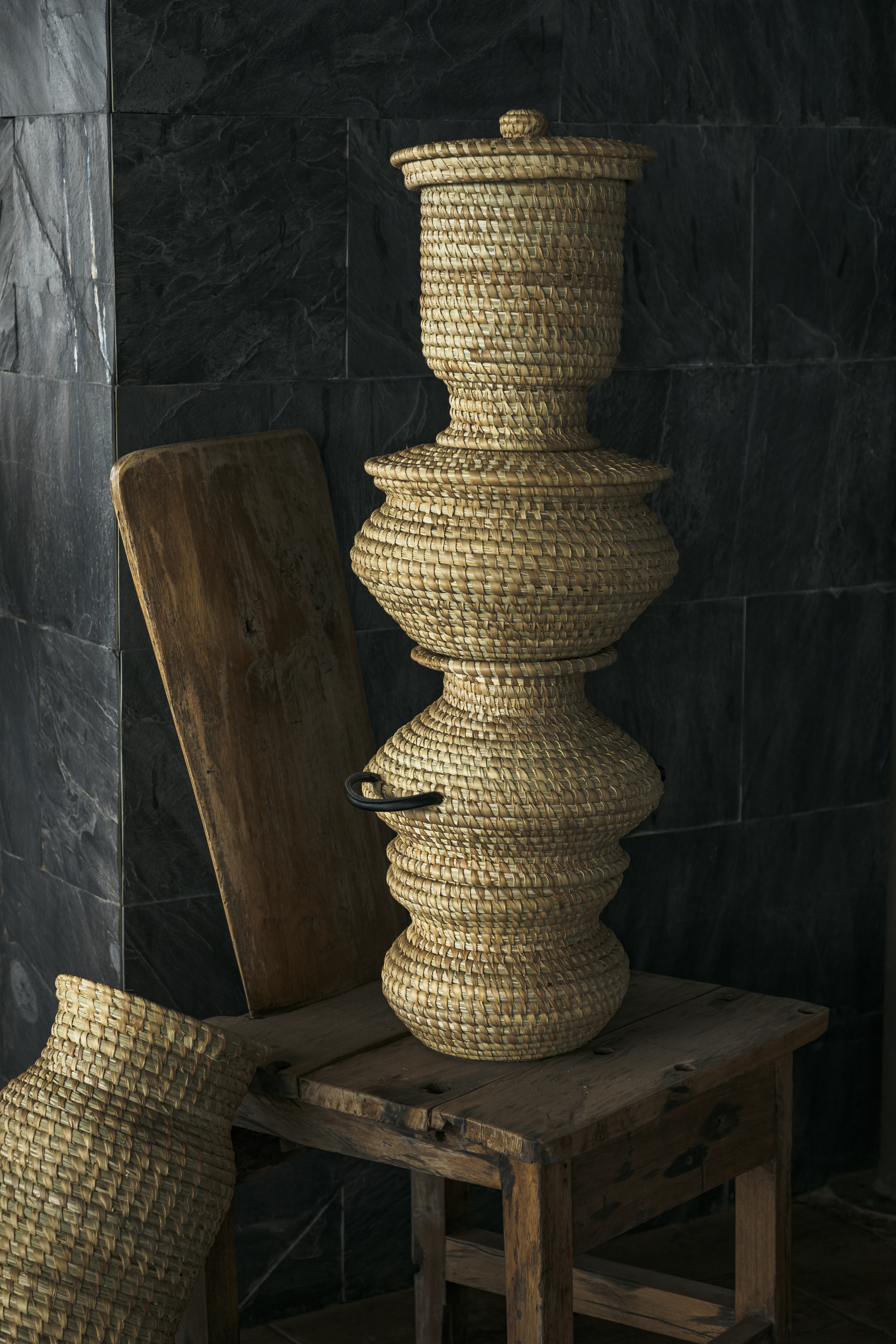 Tiered woven straw basket with lid on rustic wooden stool, dark wall.