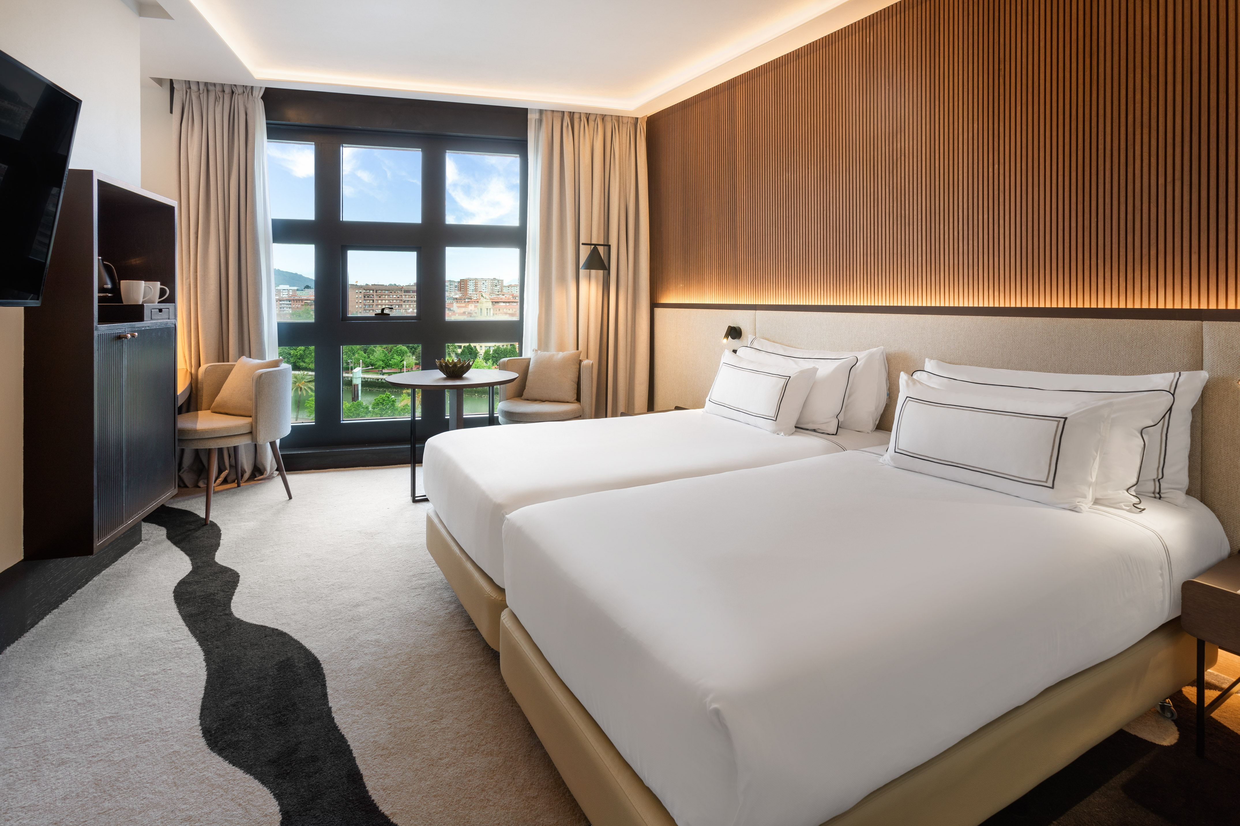 Modern hotel room with twin beds, city river view, and illuminated wooden wall.
