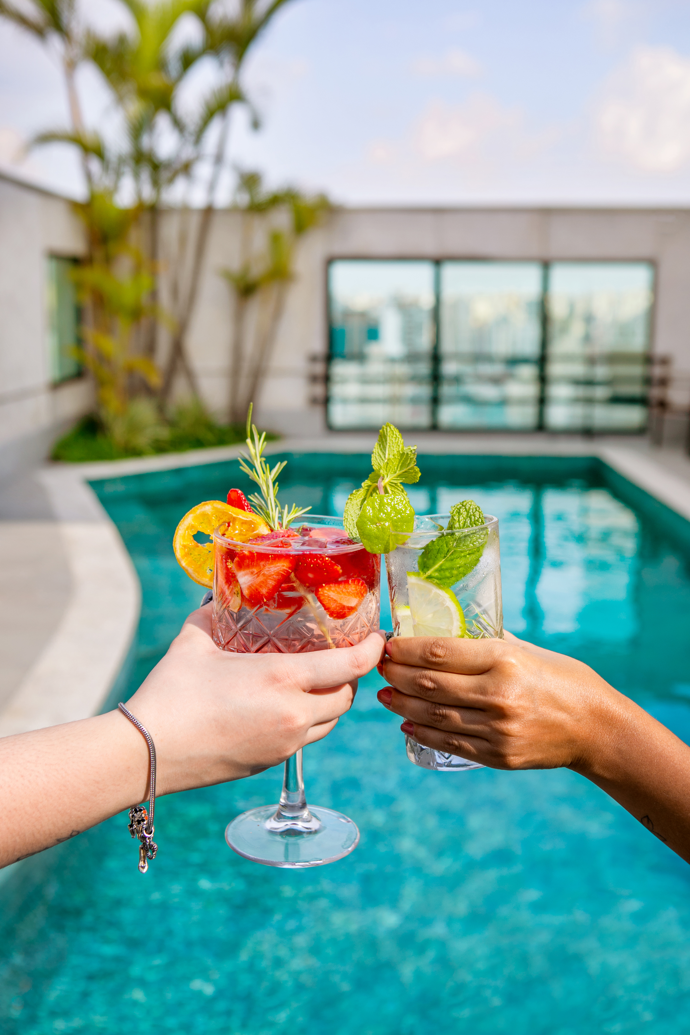 Hands toasting colorful cocktails with fruit and mint at a rooftop pool.