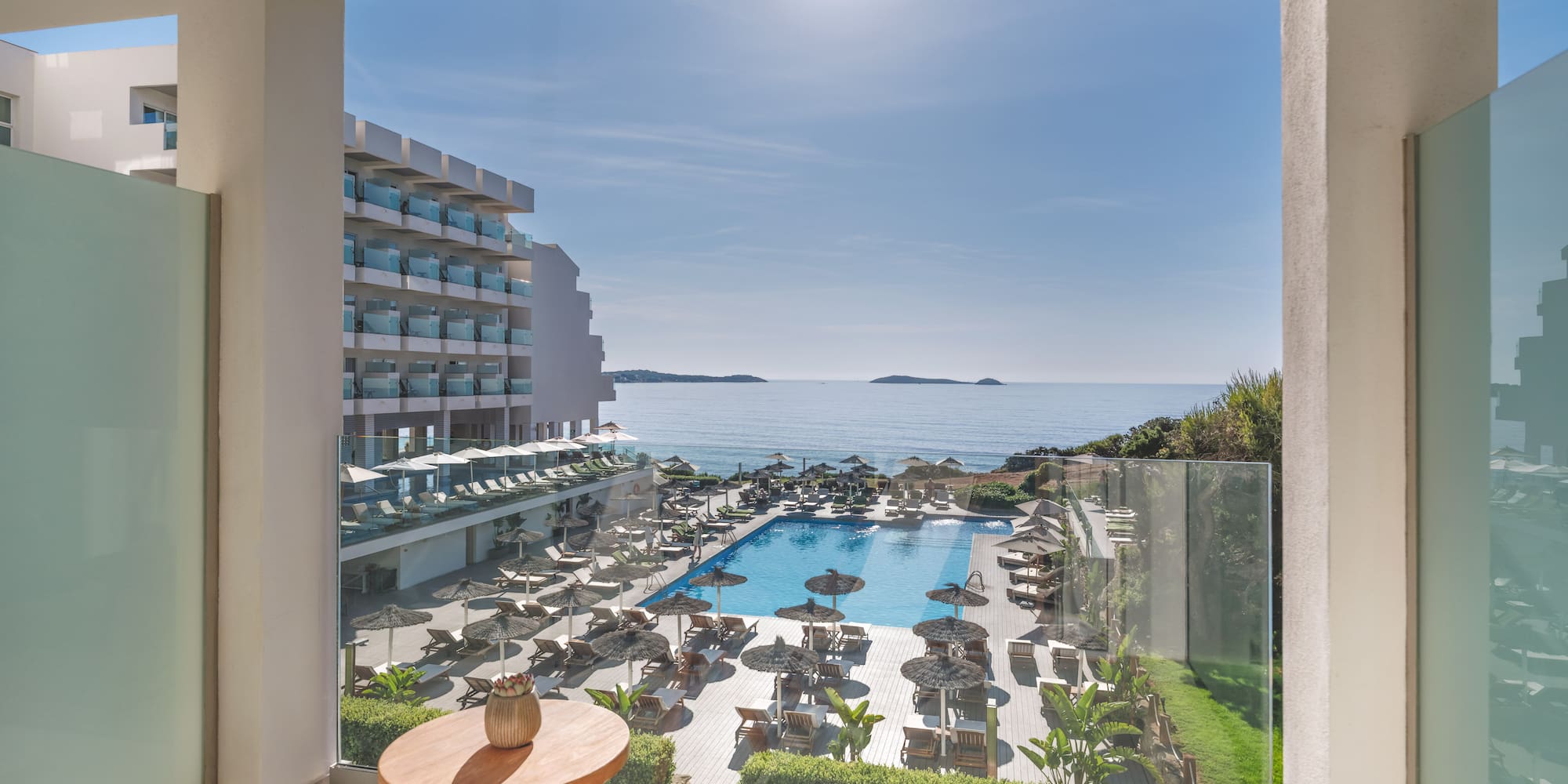 Sunny hotel balcony view of ocean, large swimming pool, and resort grounds.