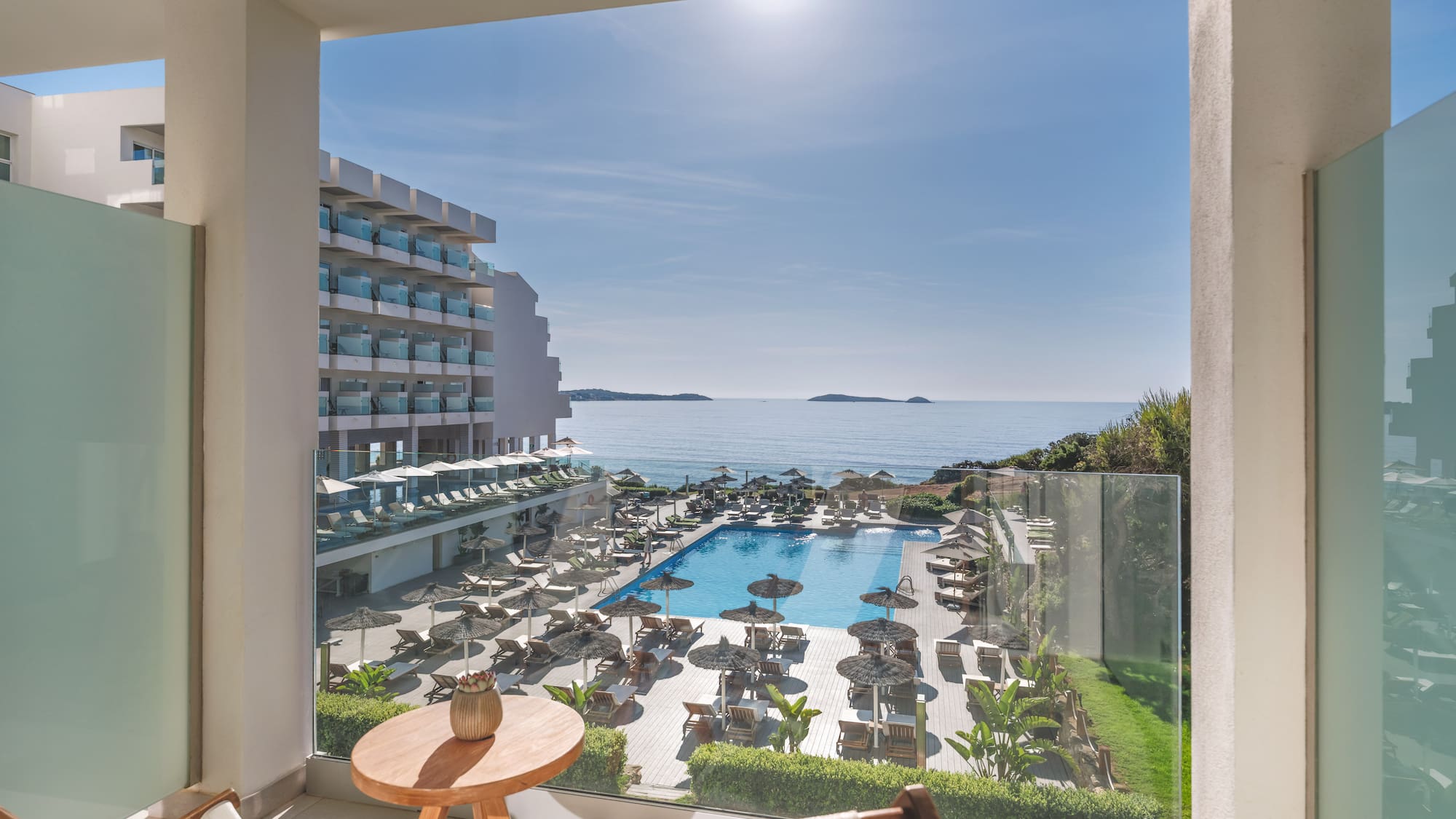 Sunny hotel balcony view of ocean, large swimming pool, and resort grounds.