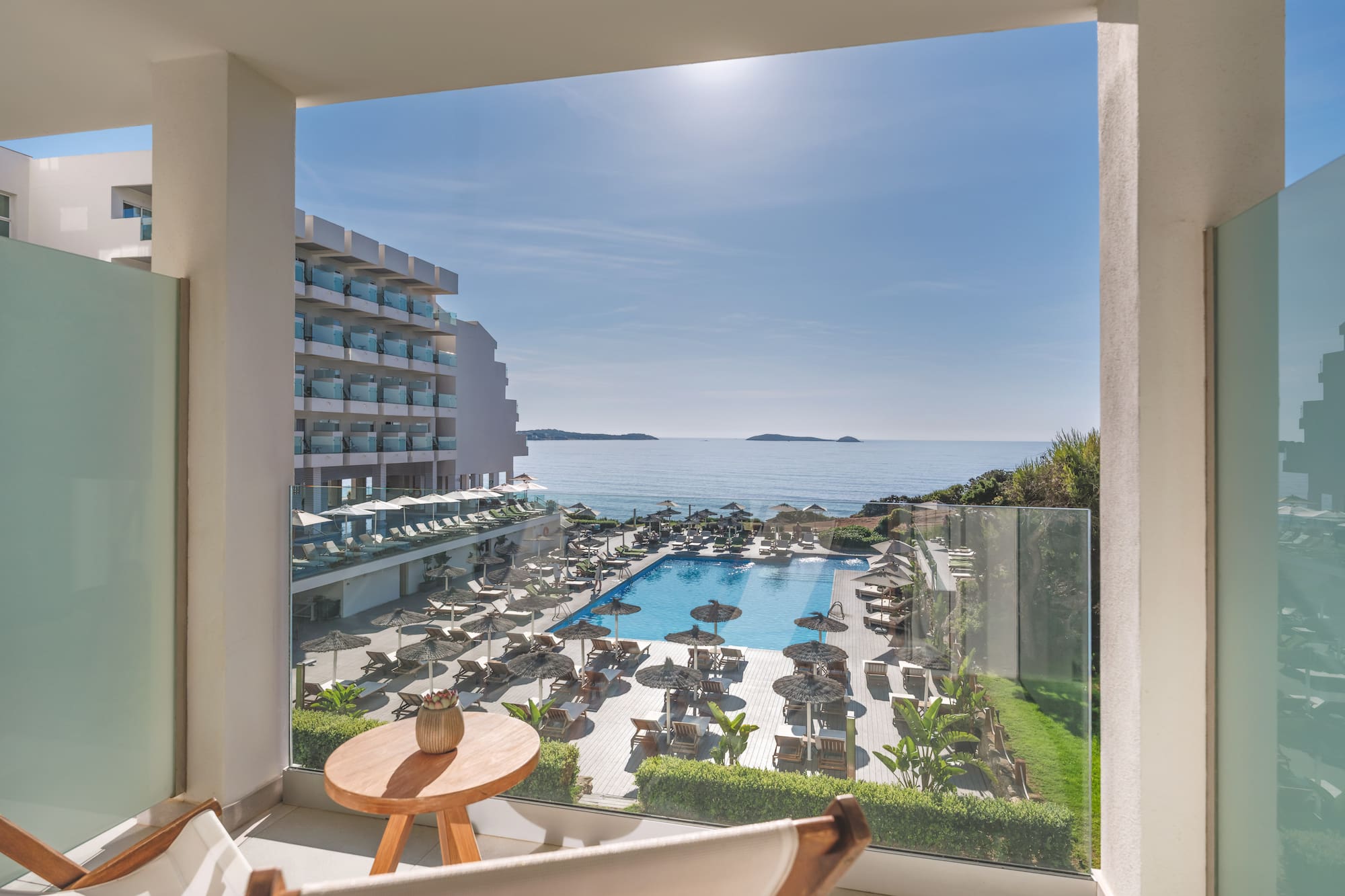 Sunny hotel balcony view of ocean, large swimming pool, and resort grounds.