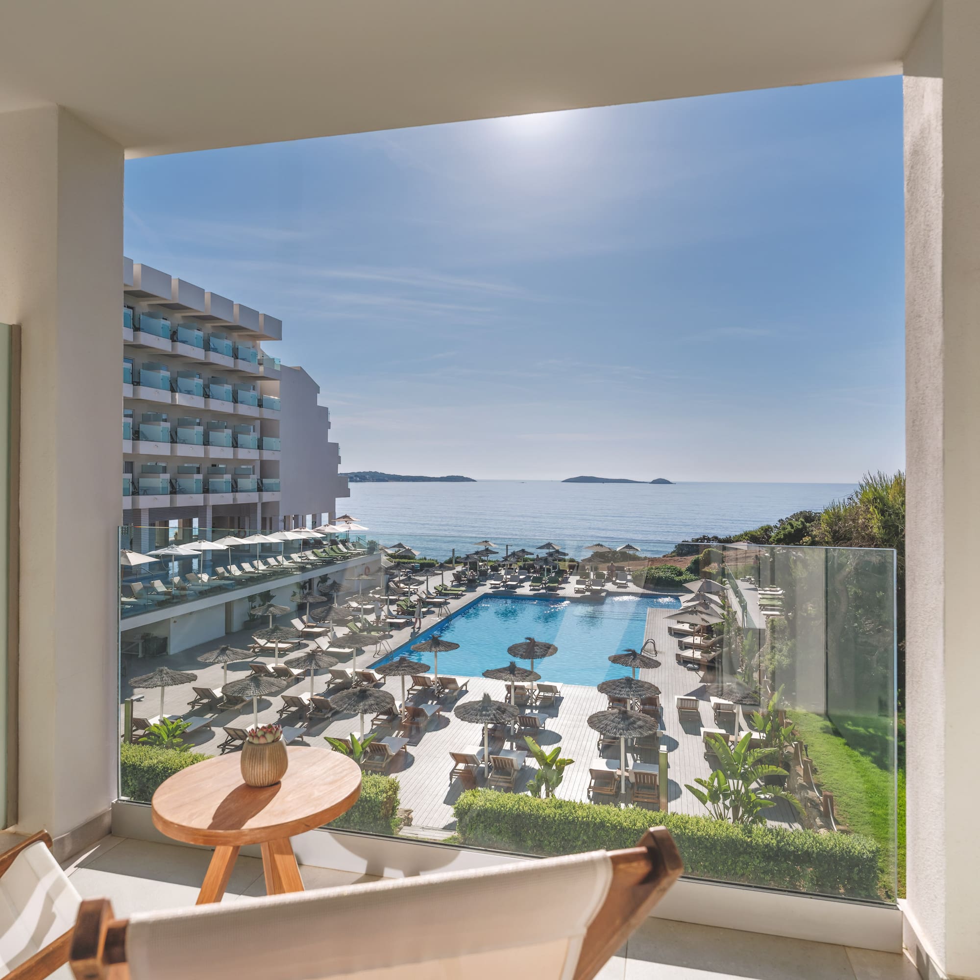 Sunny hotel balcony view of ocean, large swimming pool, and resort grounds.