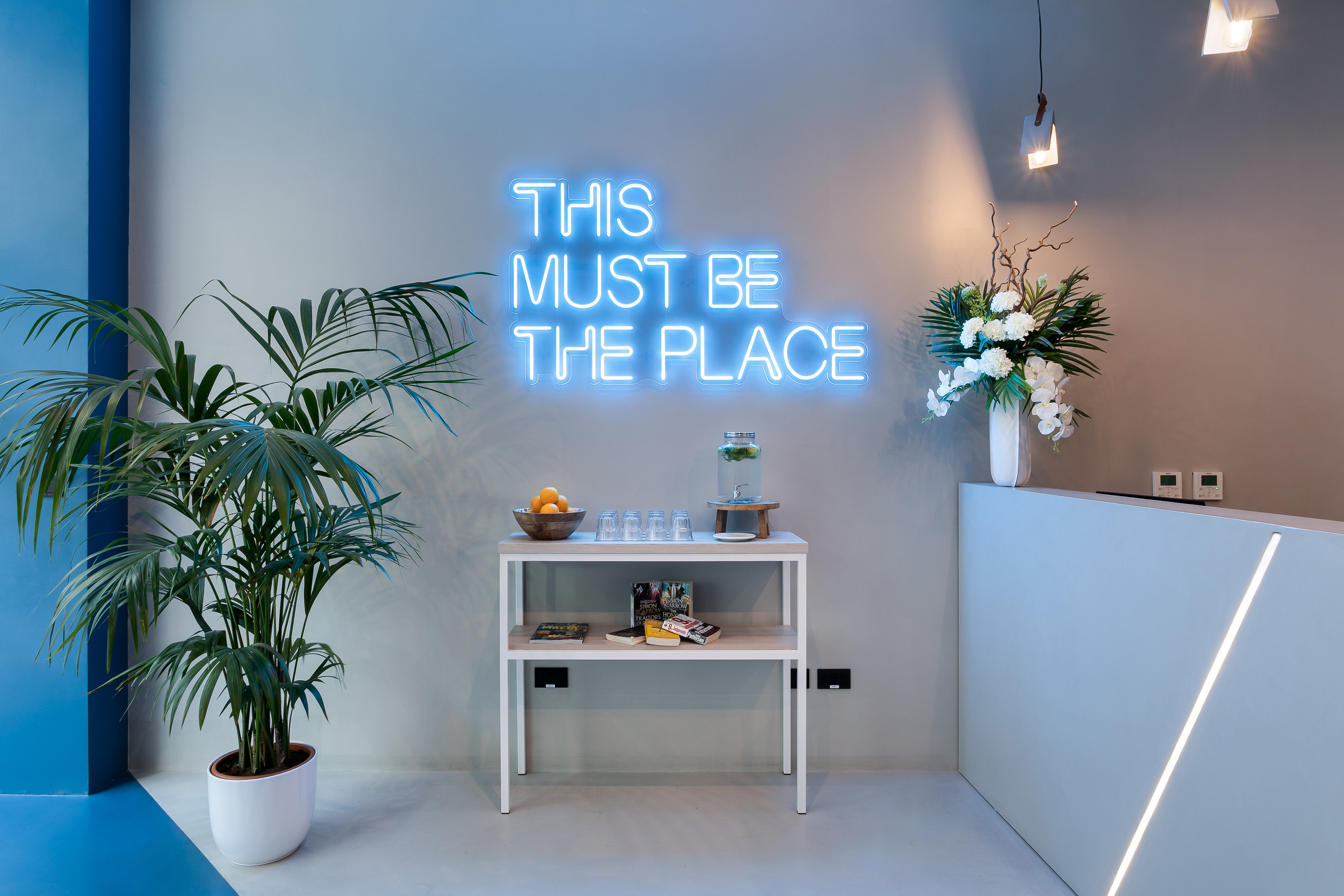 Modern reception with blue neon sign, palm plant, and sleek white desk.