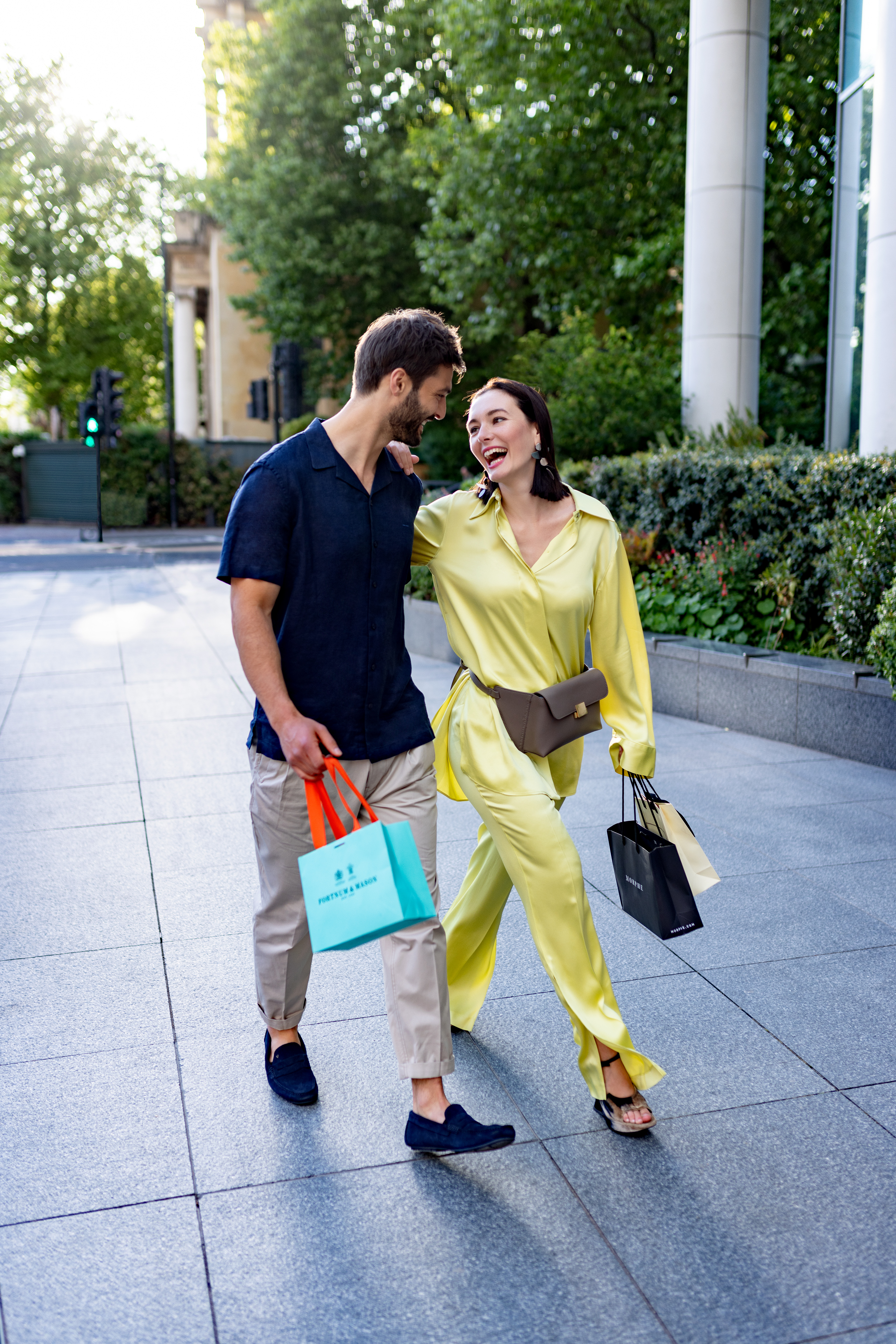 A man and a woman stroll along a sidewalk, each carrying bags.