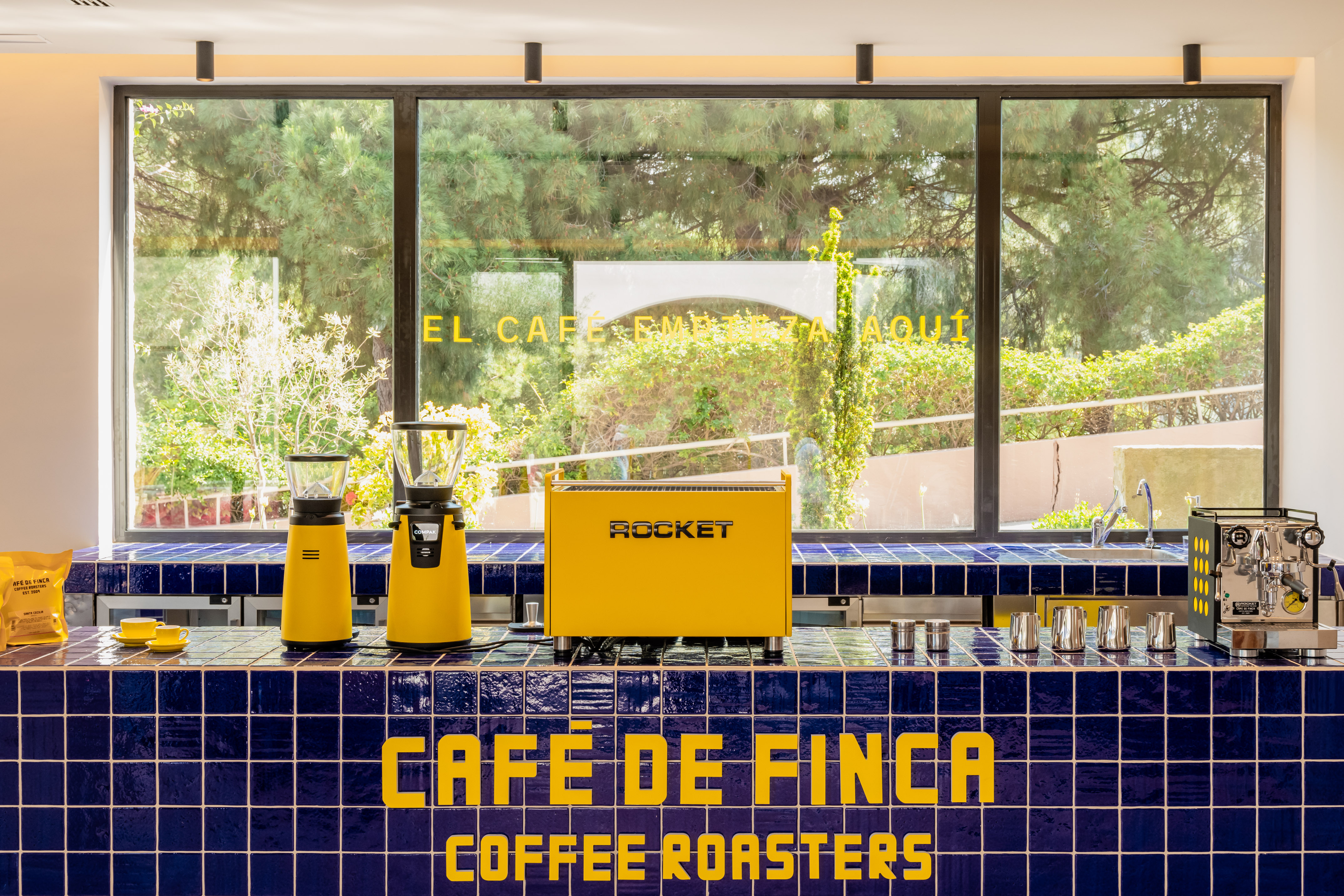 Cafe de Finca coffee bar: yellow espresso machine, grinders, blue tiles.