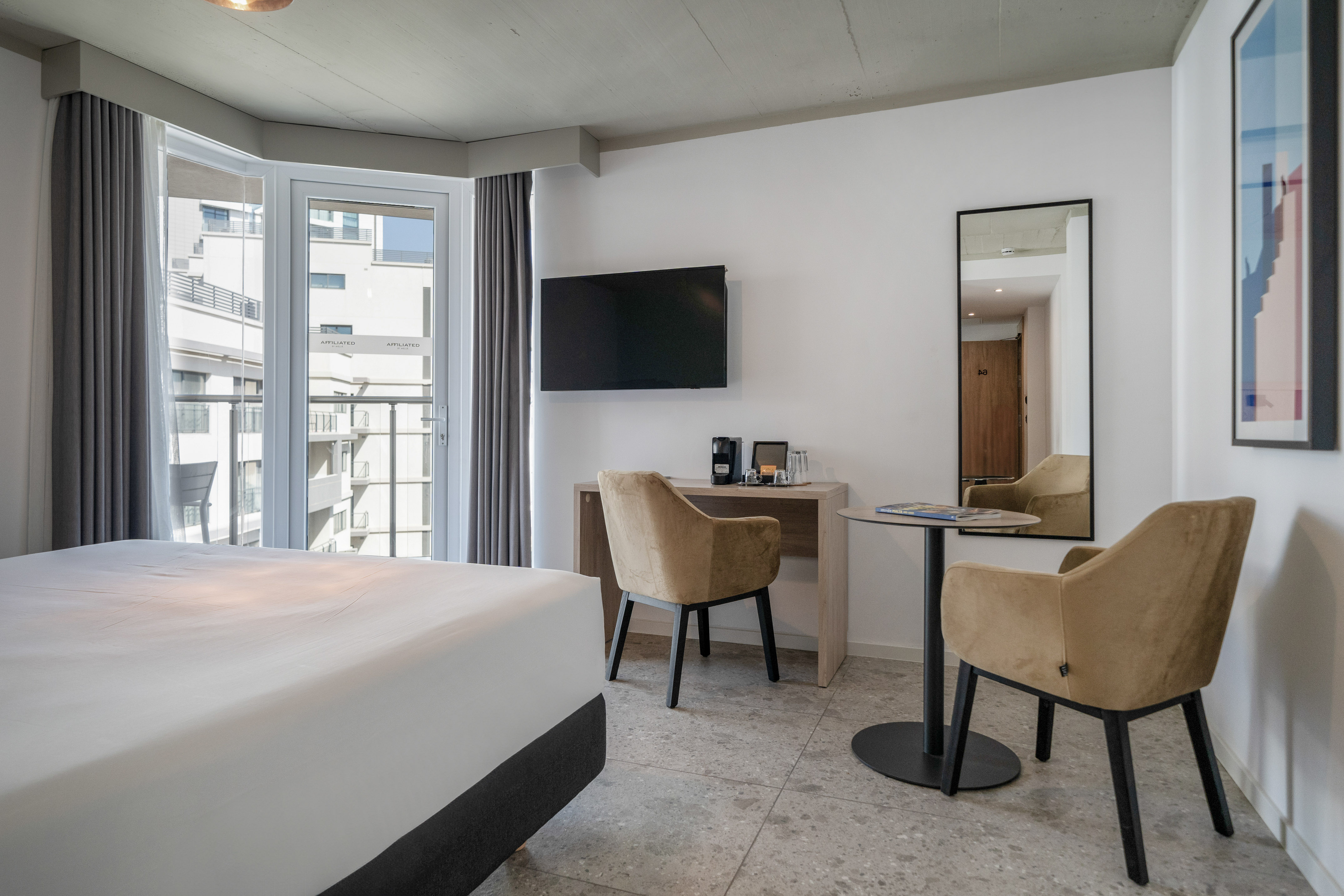 Modern hotel room with balcony, large bed, TV, work desk, and two chairs.