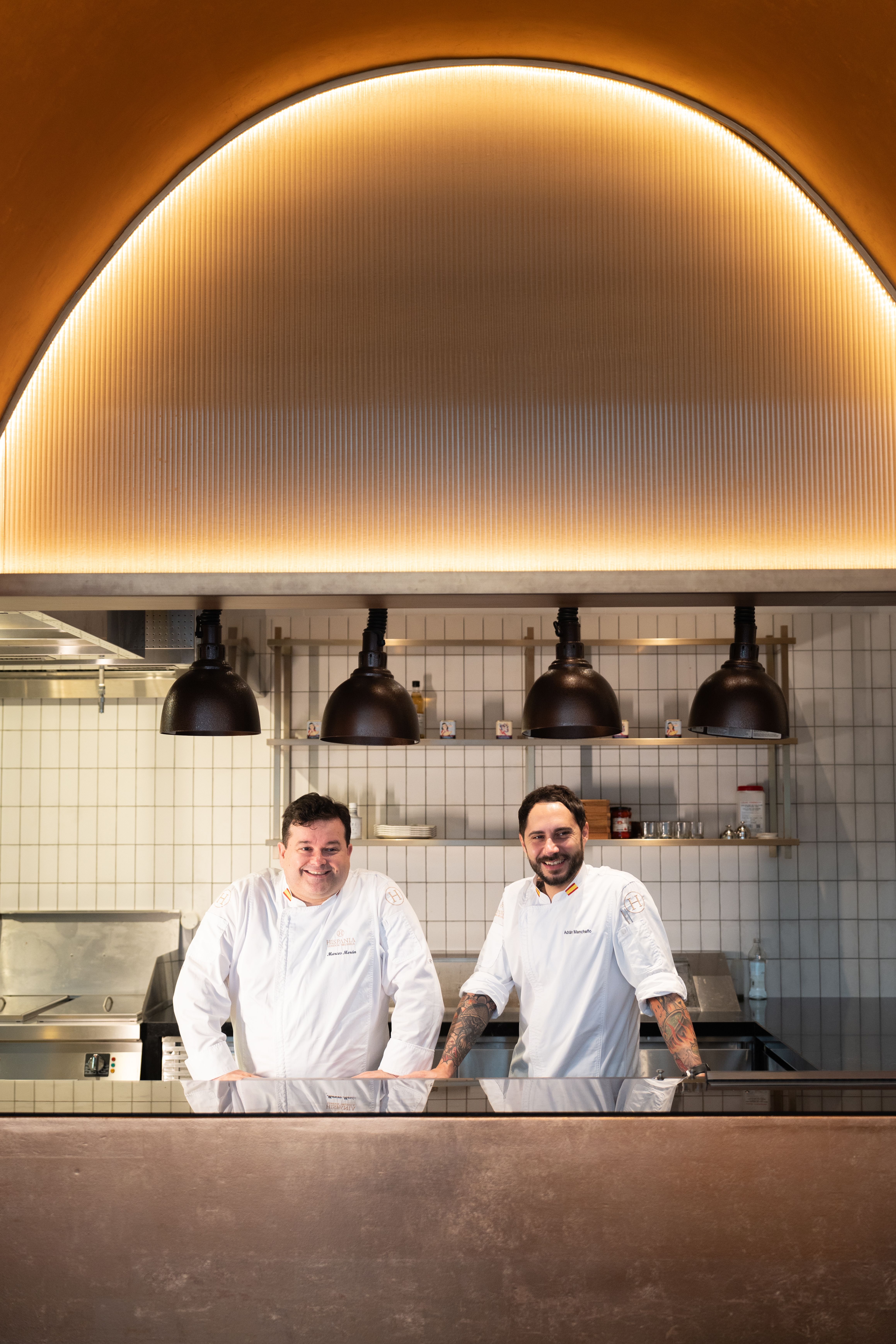 Smiling chefs Marcos Morán and Adrian Mancheño in a professional restaurant kitchen.