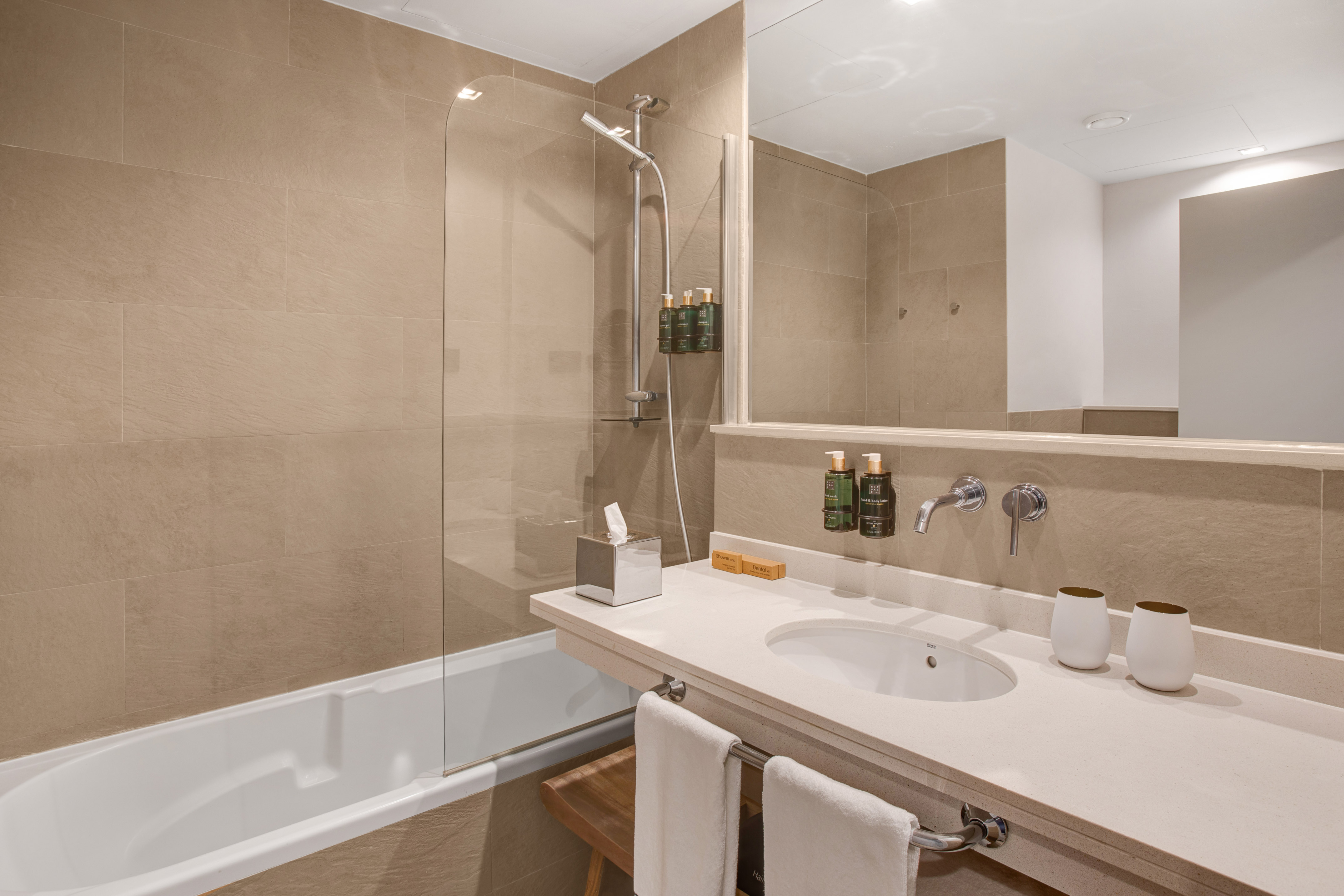 Modern hotel bathroom with beige tiles, white tub, vanity, and mirror.