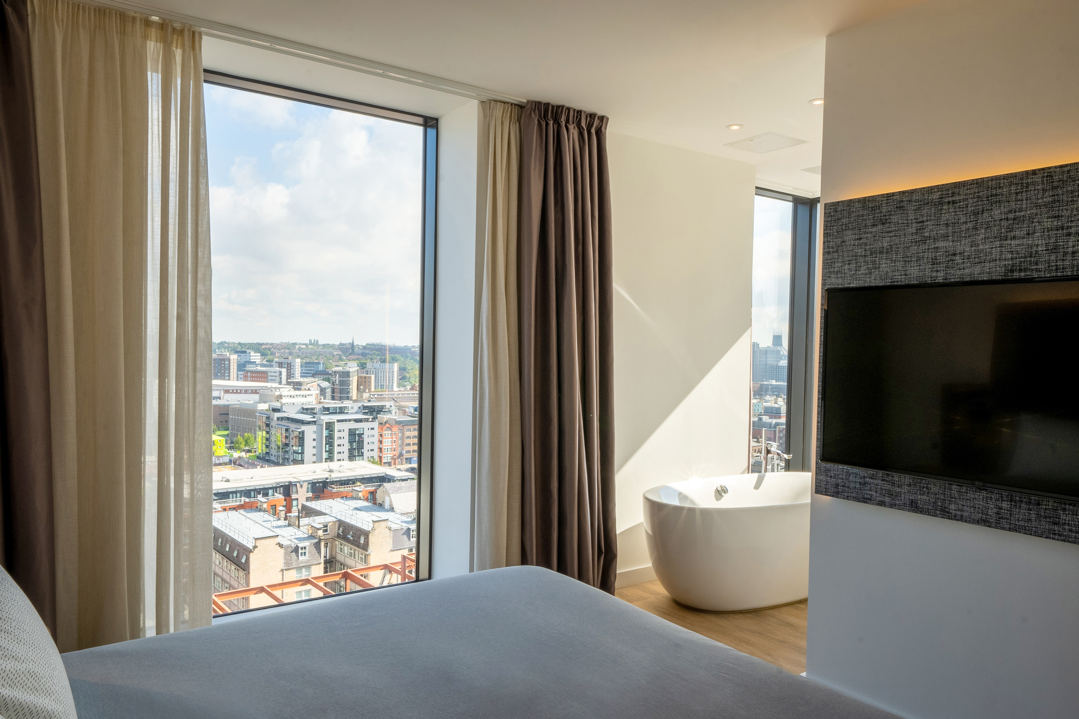 Modern hotel room with panoramic city view, bed, and freestanding bathtub.
