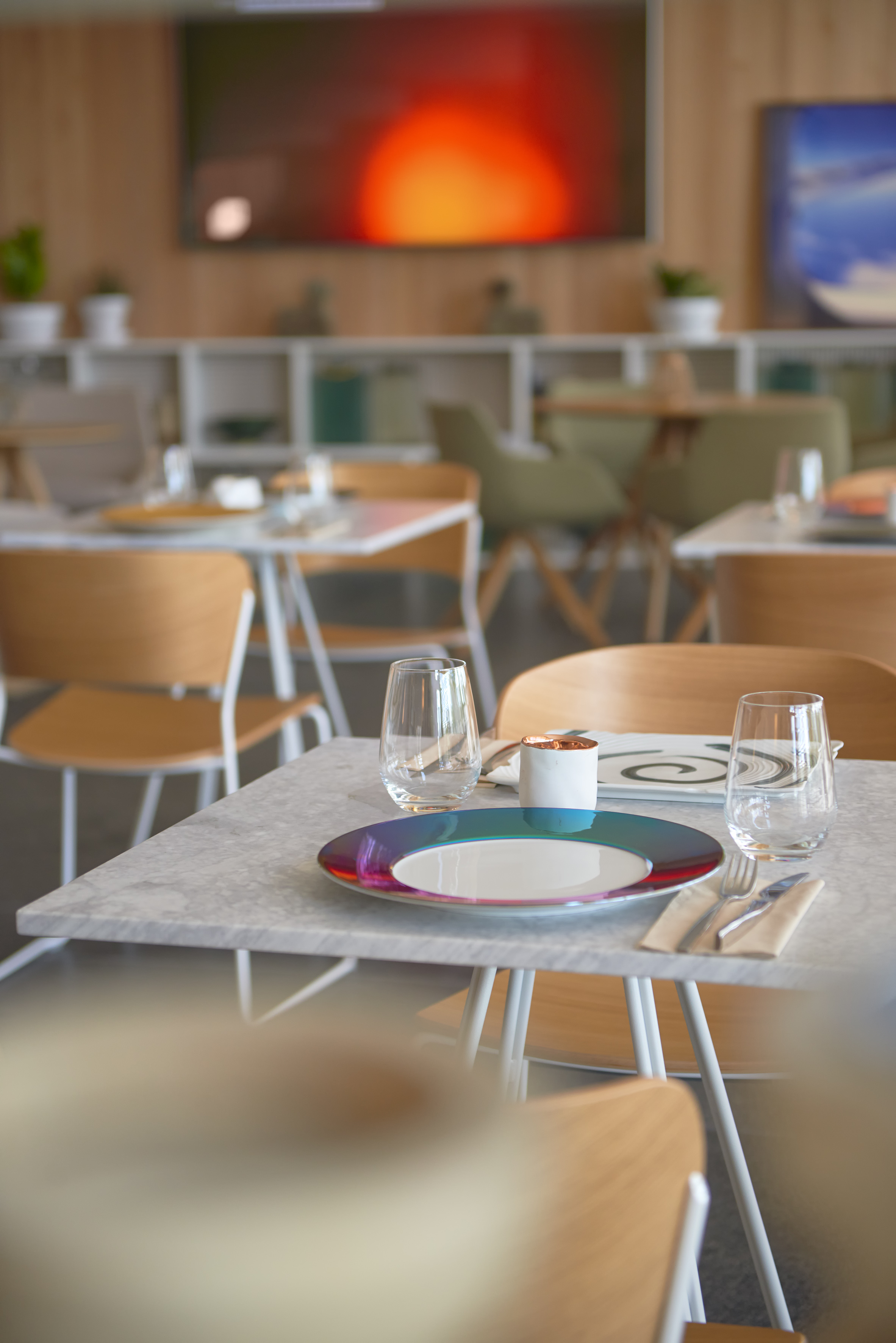 Modern restaurant interior with colorful table setting, wooden chairs, and a large screen.