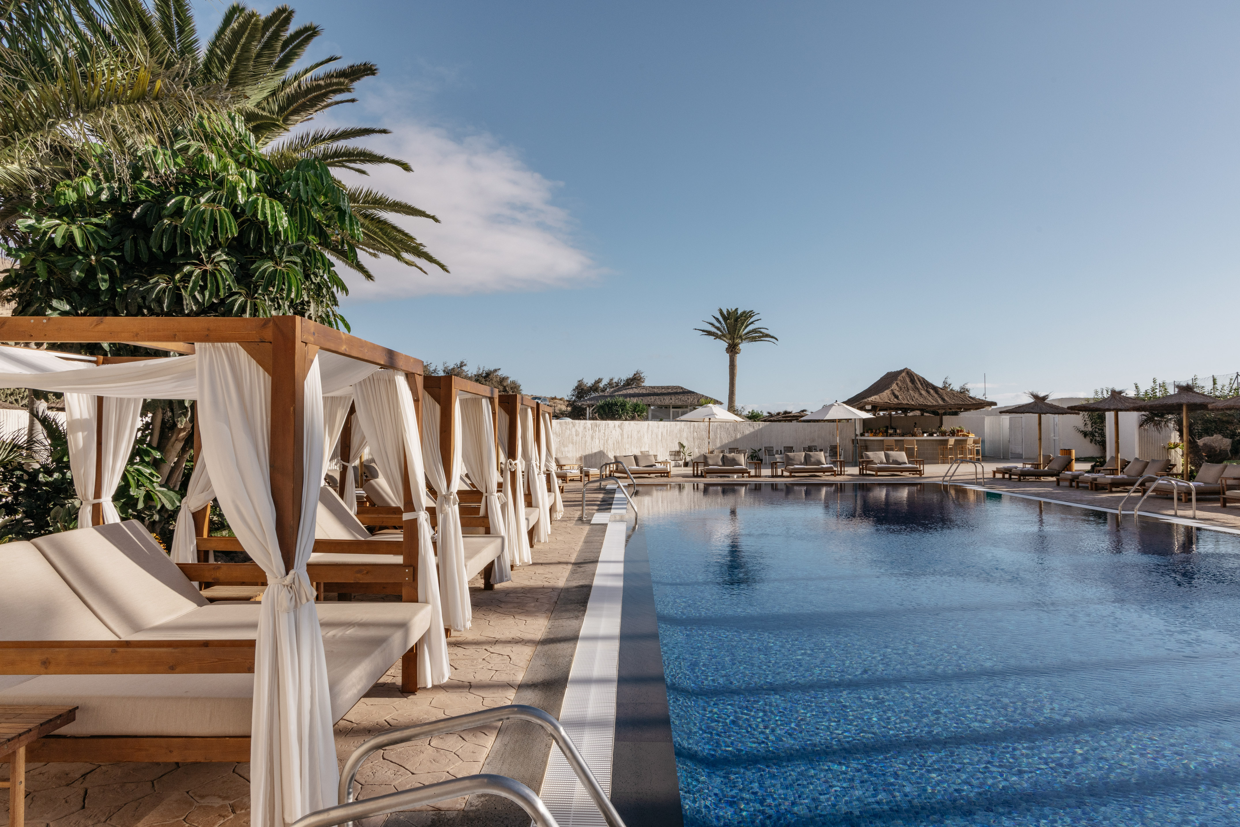 Sunny resort pool with a row of wooden cabanas, palm trees, and a bar.