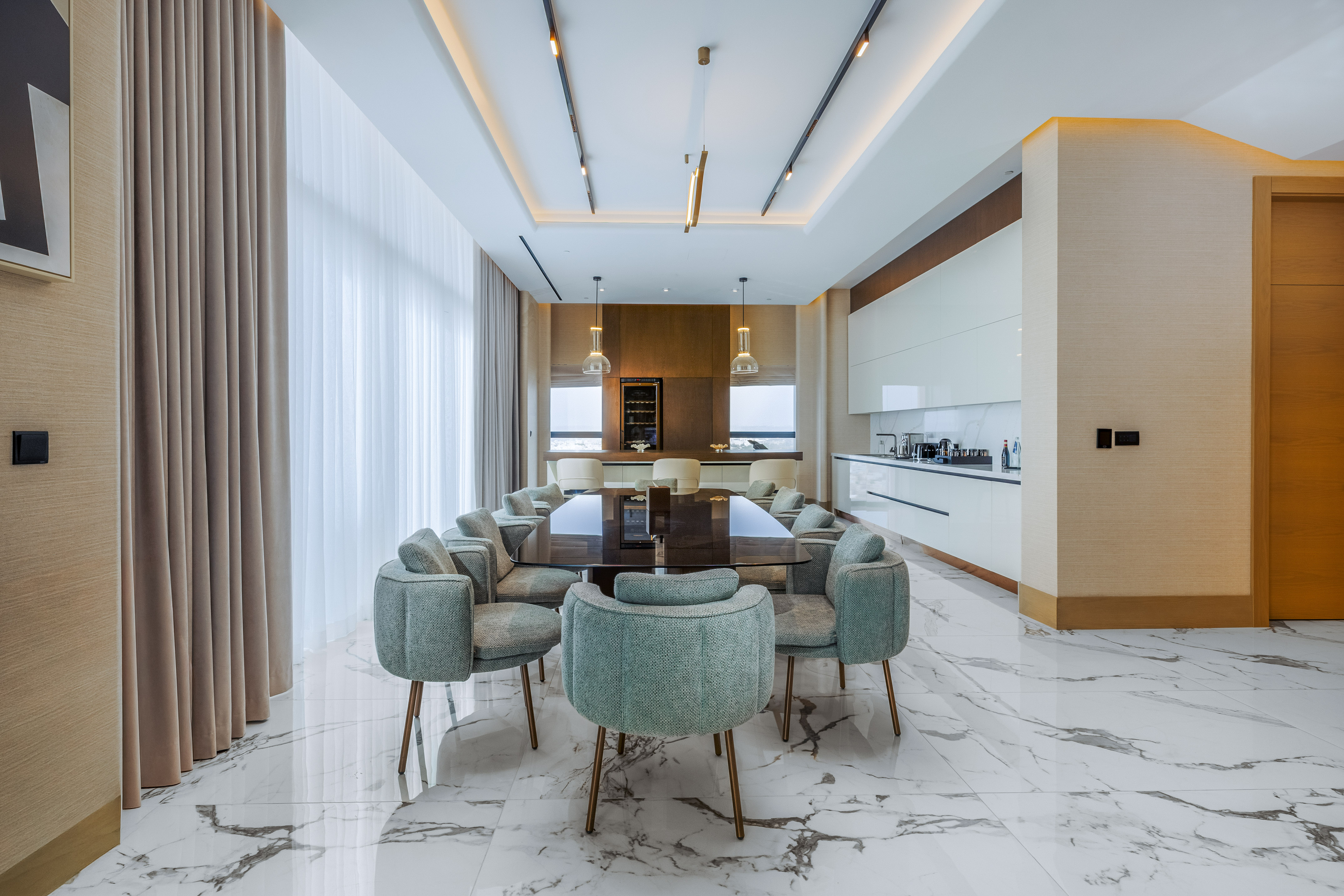 Modern luxury dining room with marble floor, large table, green chairs, and integrated kitchen.
