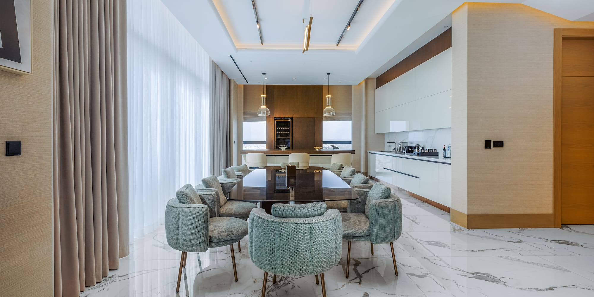 Modern luxury dining room with marble floor, large table, green chairs, and integrated kitchen.