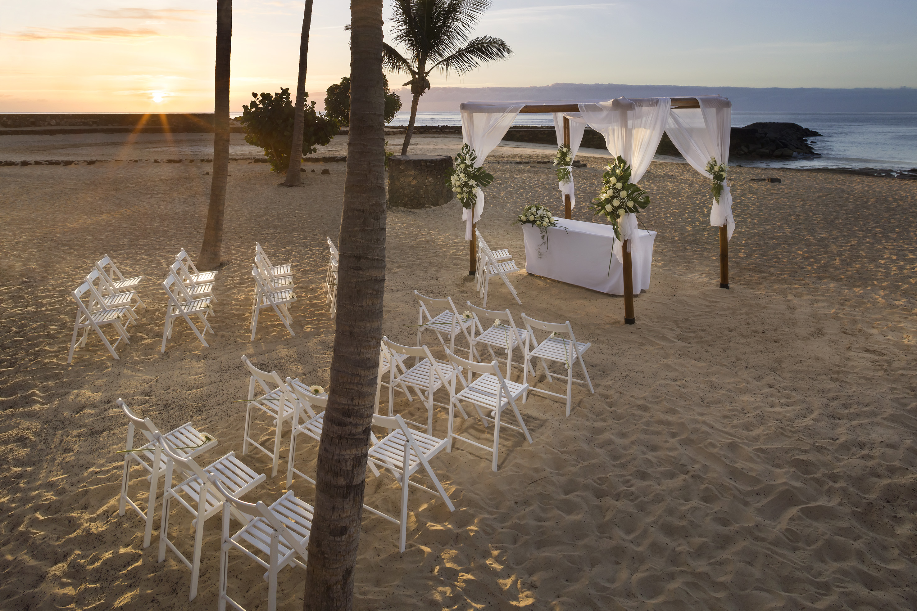 Beach wedding ceremony with white chairs, canopy, ocean, and sunrise.
