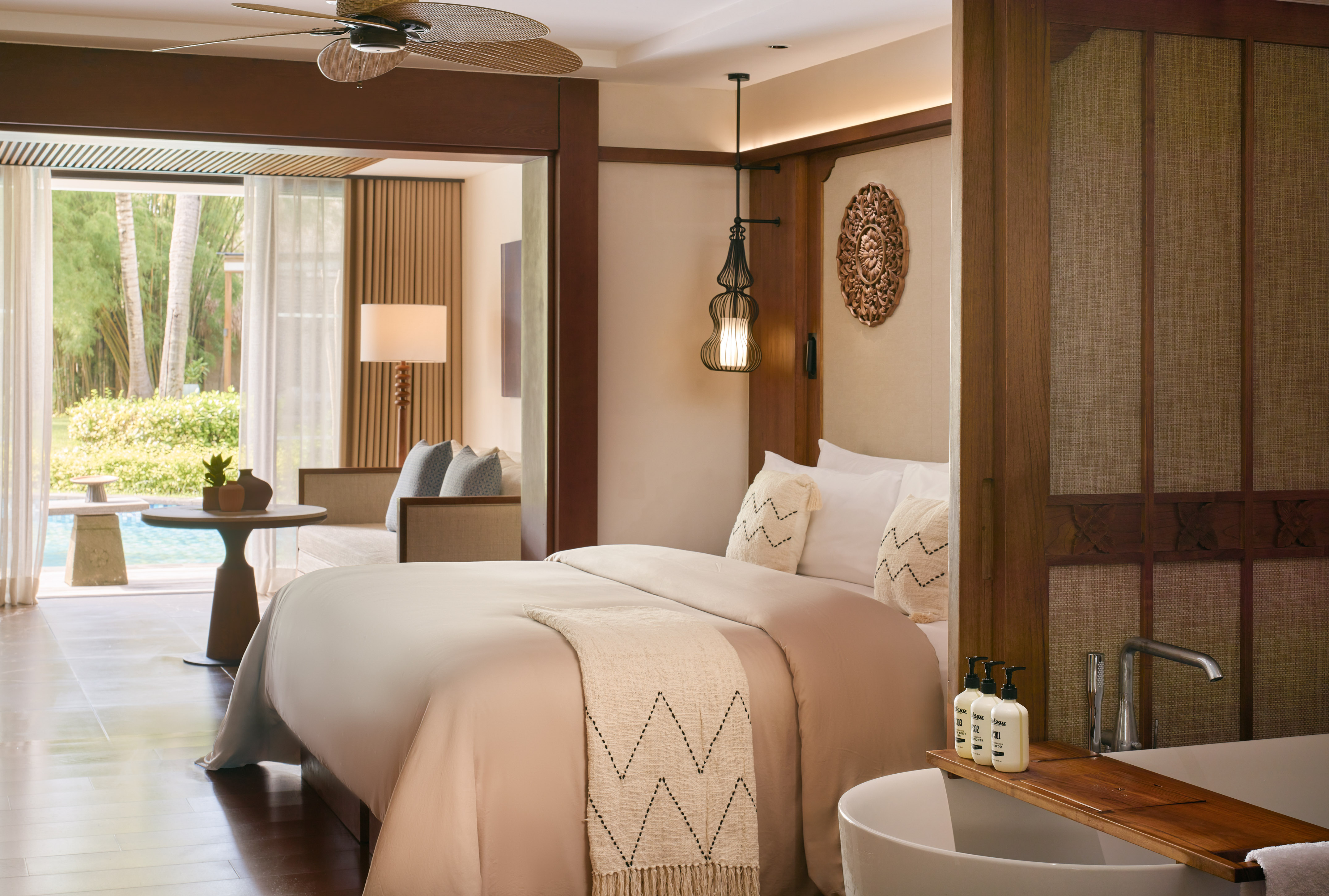 Luxury hotel suite with bed, freestanding bathtub, and lush pool garden view.