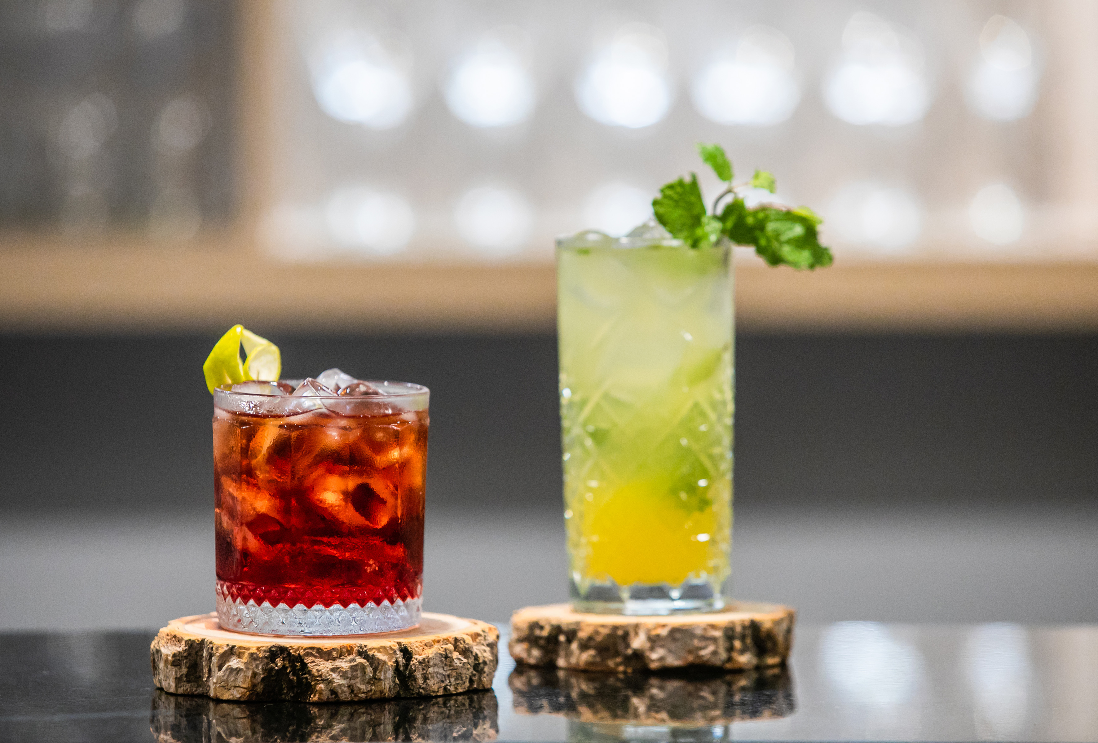 Red cocktail with lemon, green cocktail with mint, on wooden coasters.