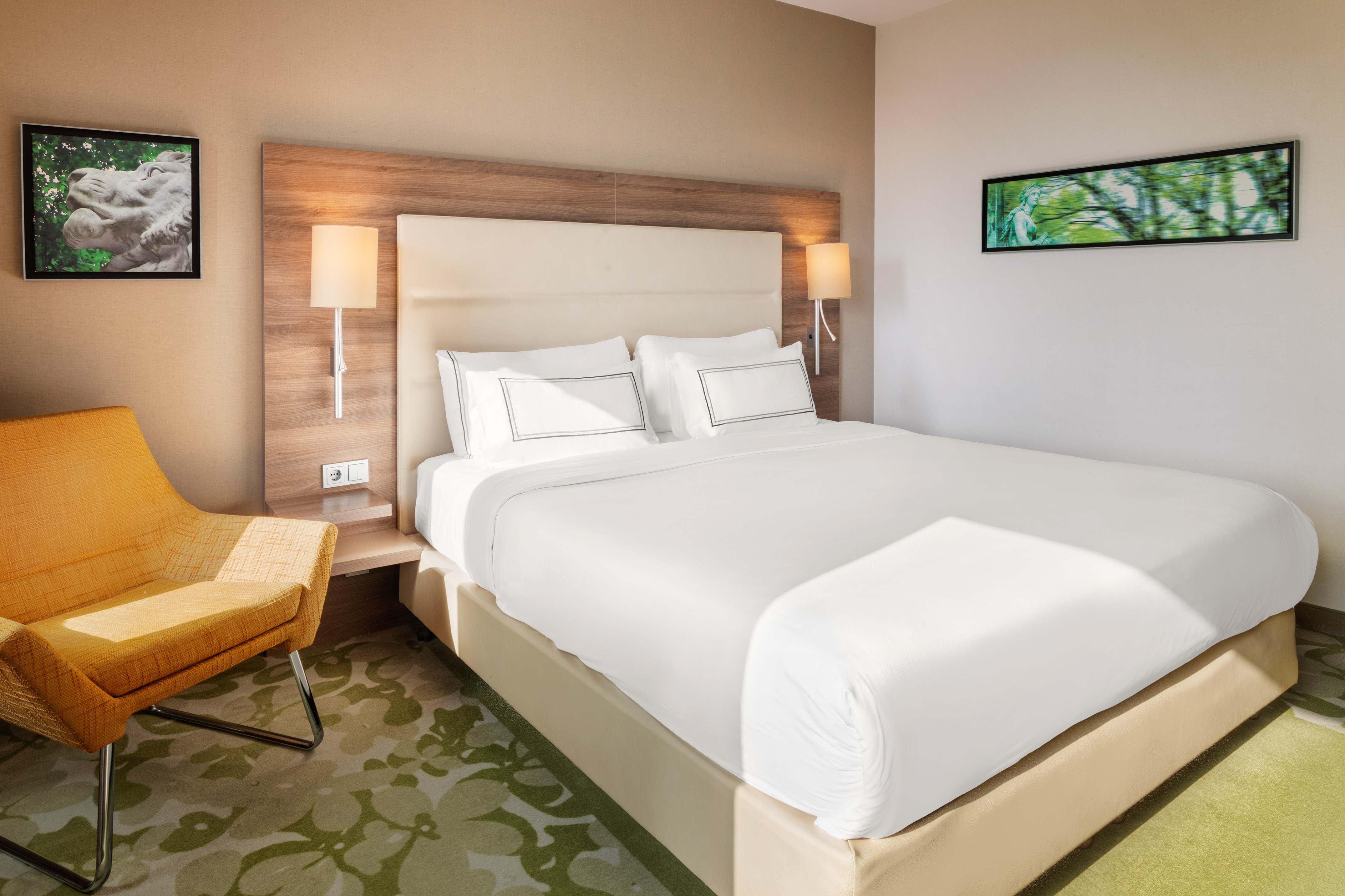 Modern hotel bedroom with king bed, white linens, orange armchair, and green carpet.