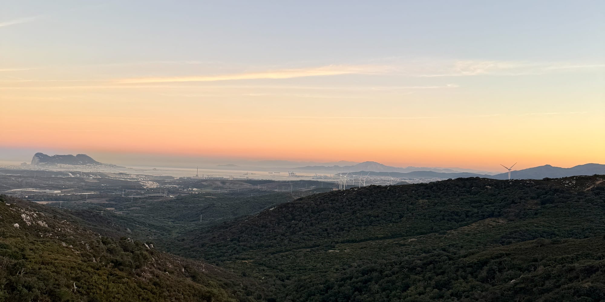 Sunrise over green valleys, distant Rock of Gibraltar, city, and wind turbines.