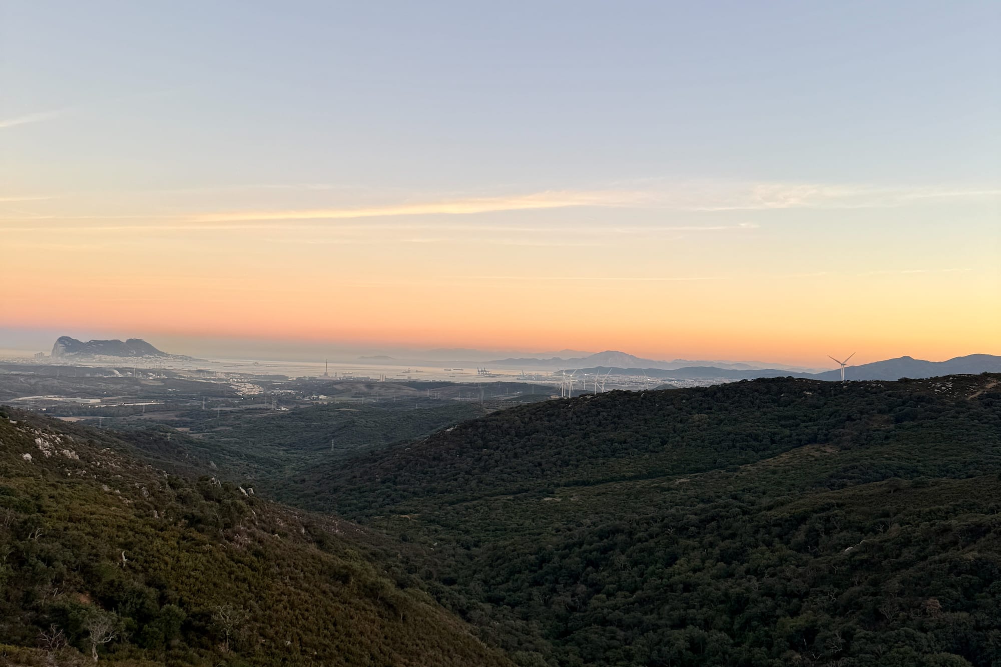 Sunrise over green valleys, distant Rock of Gibraltar, city, and wind turbines.