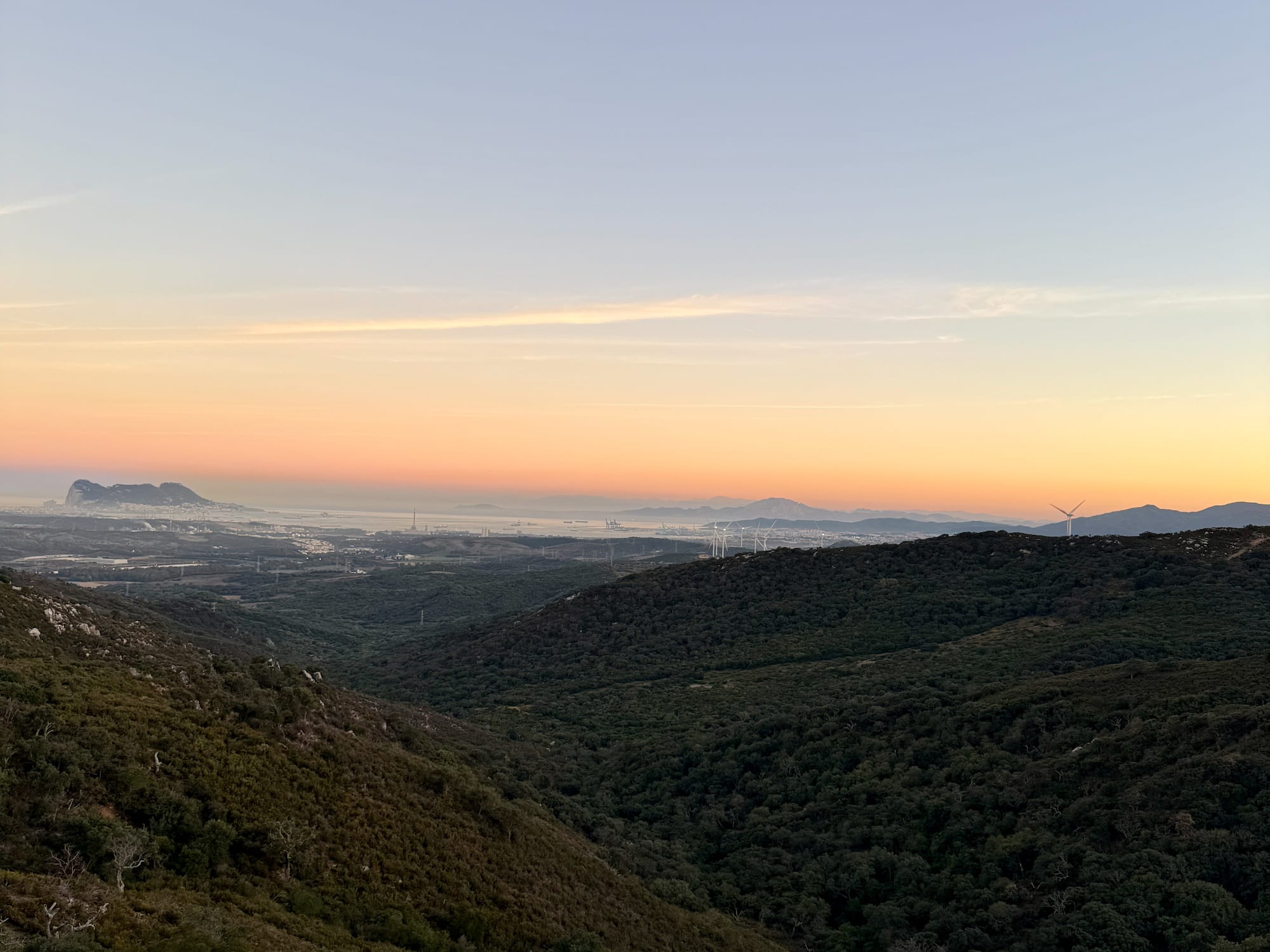 Sunrise over green valleys, distant Rock of Gibraltar, city, and wind turbines.