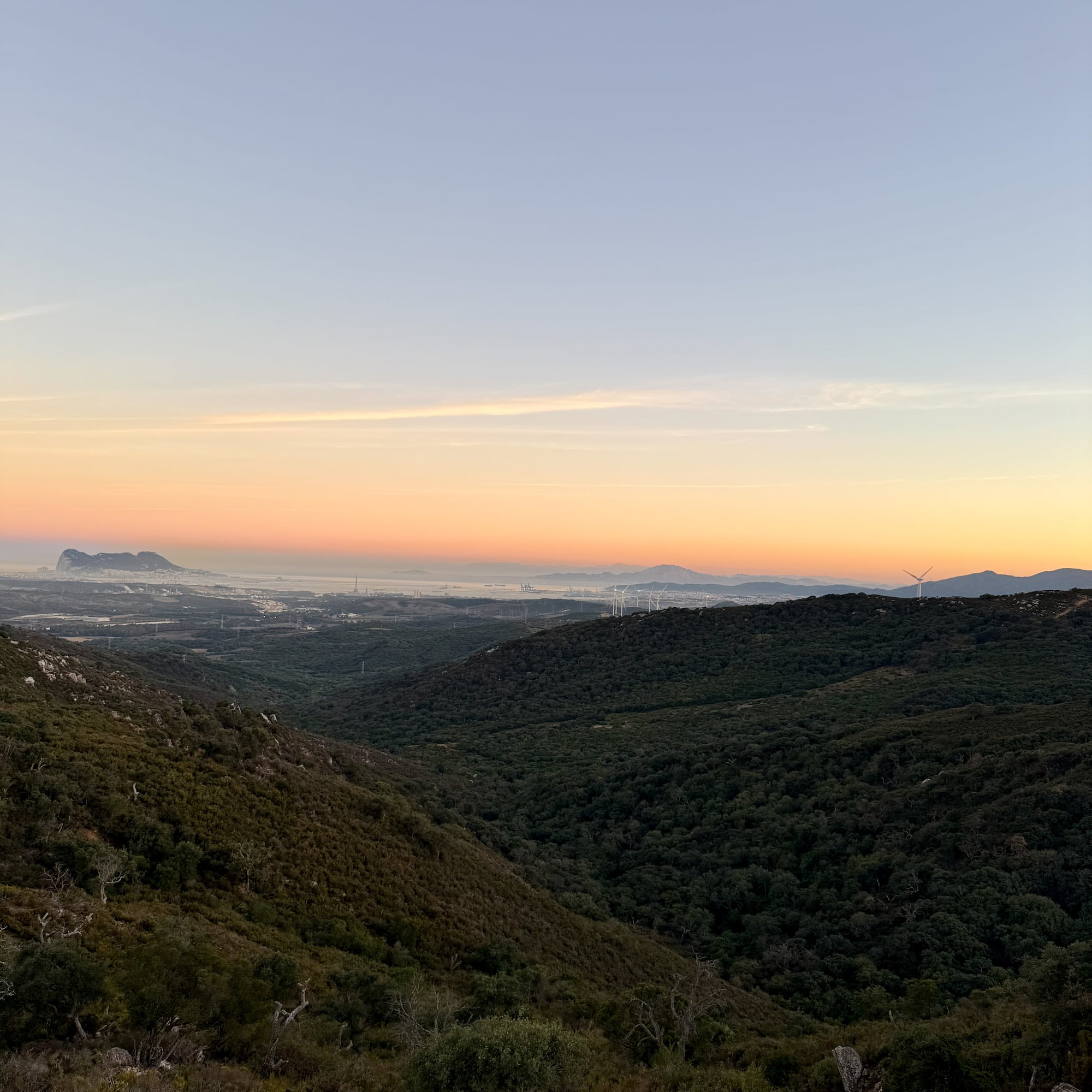 Sunrise over green valleys, distant Rock of Gibraltar, city, and wind turbines.