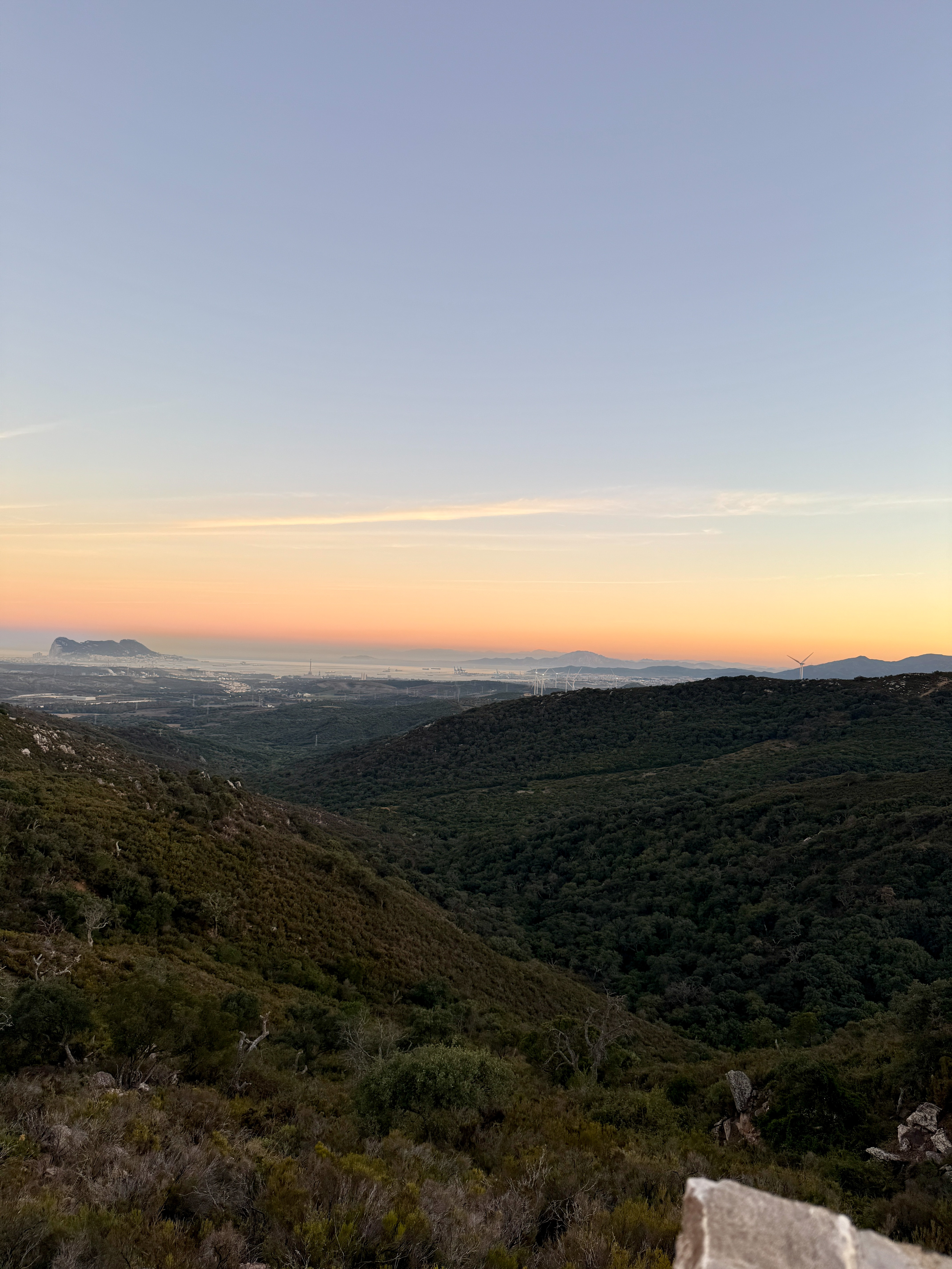 Sunrise over green valleys, distant Rock of Gibraltar, city, and wind turbines.