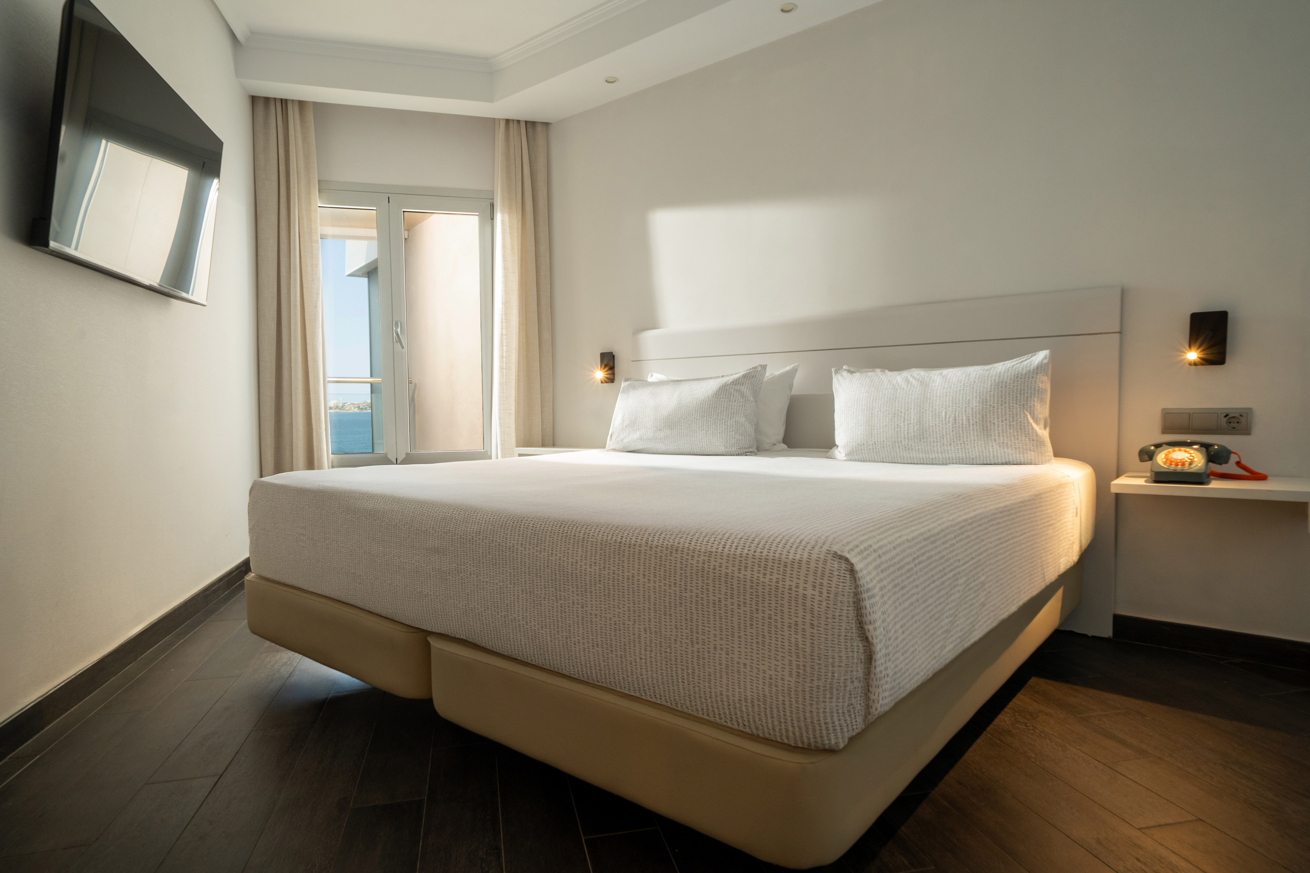Modern hotel bedroom with large bed, sea view, wall TV, and dark wood floor.