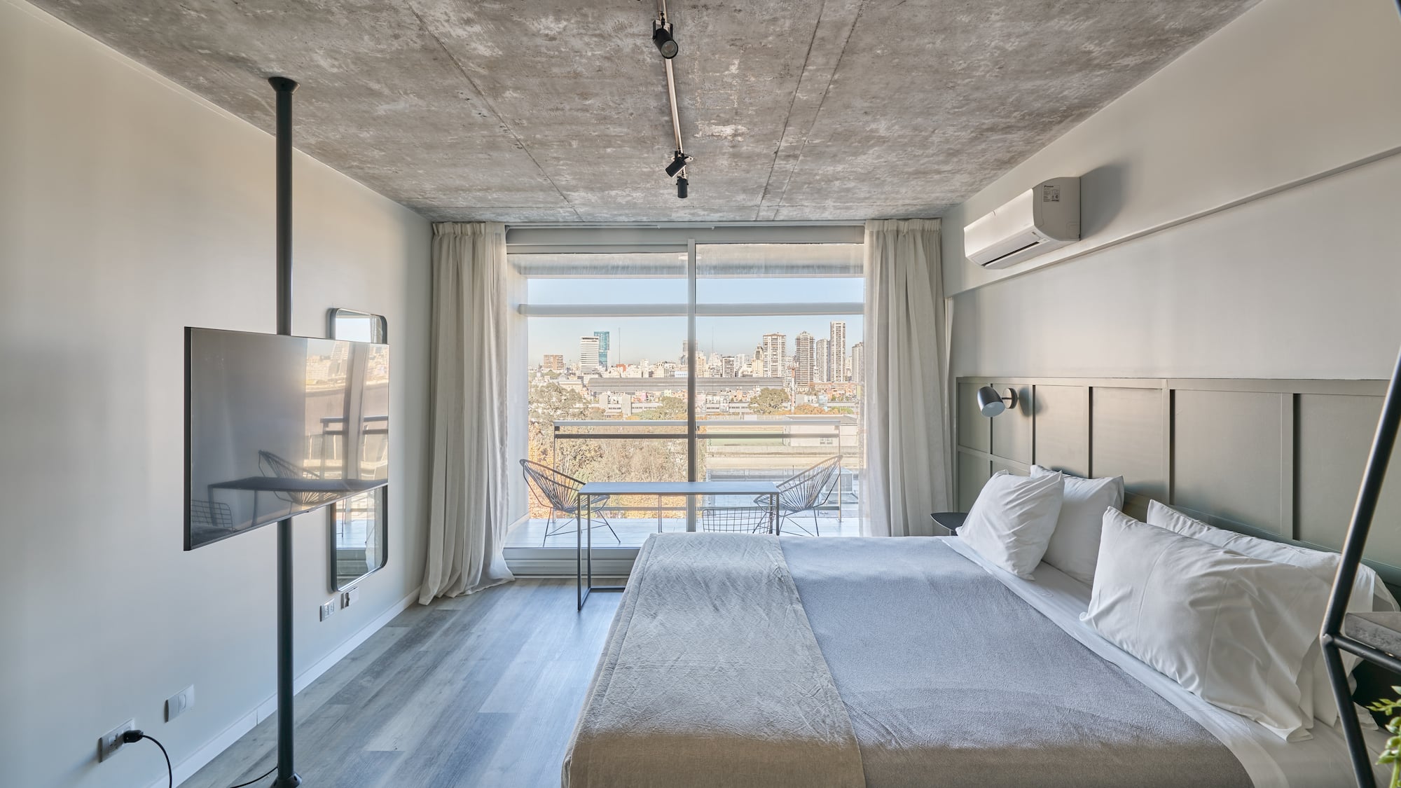 Modern bedroom with city view balcony, concrete ceiling, and king bed.