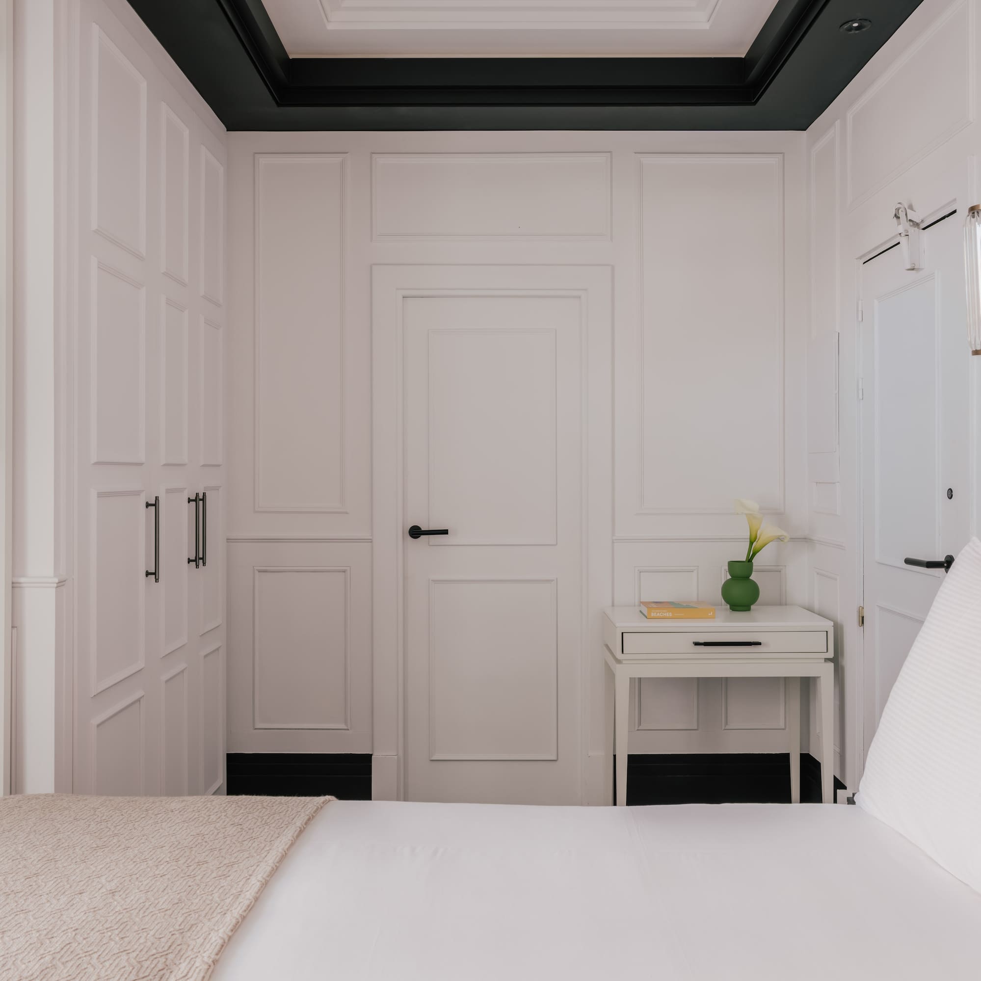 Modern white bedroom with paneled walls, bed, TV, nightstand, and wall sconce.