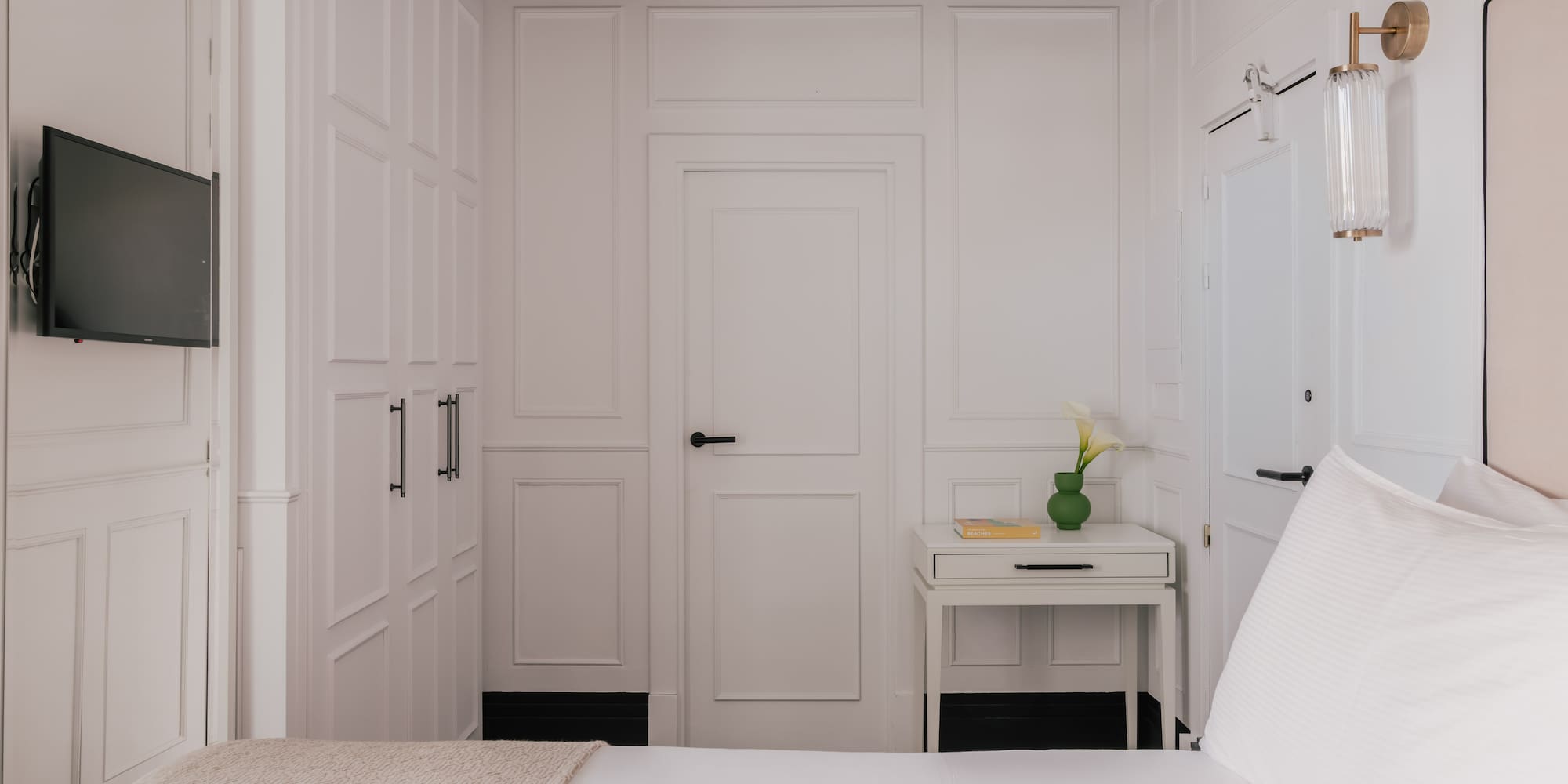 Modern white bedroom with paneled walls, bed, TV, nightstand, and wall sconce.