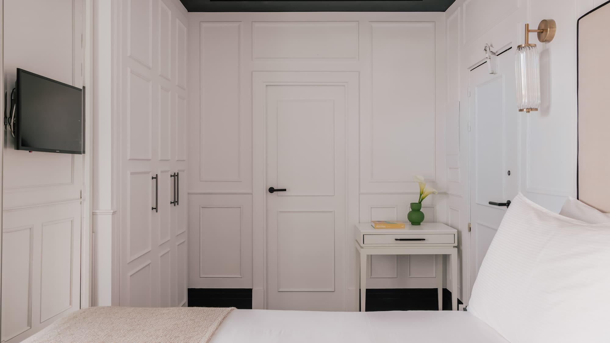 Modern white bedroom with wainscoting, black trim, bed, TV, and nightstand.