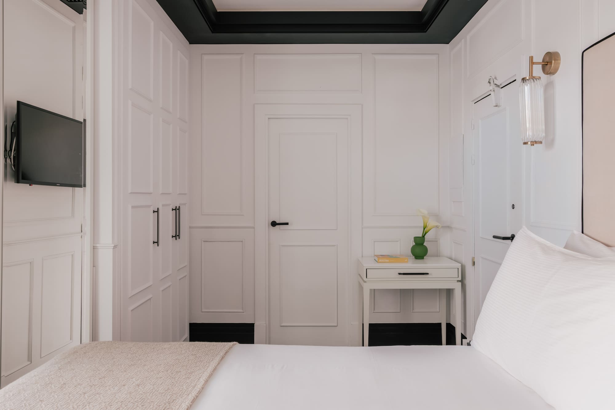 Modern white bedroom with paneled walls, bed, TV, nightstand, and wall sconce.