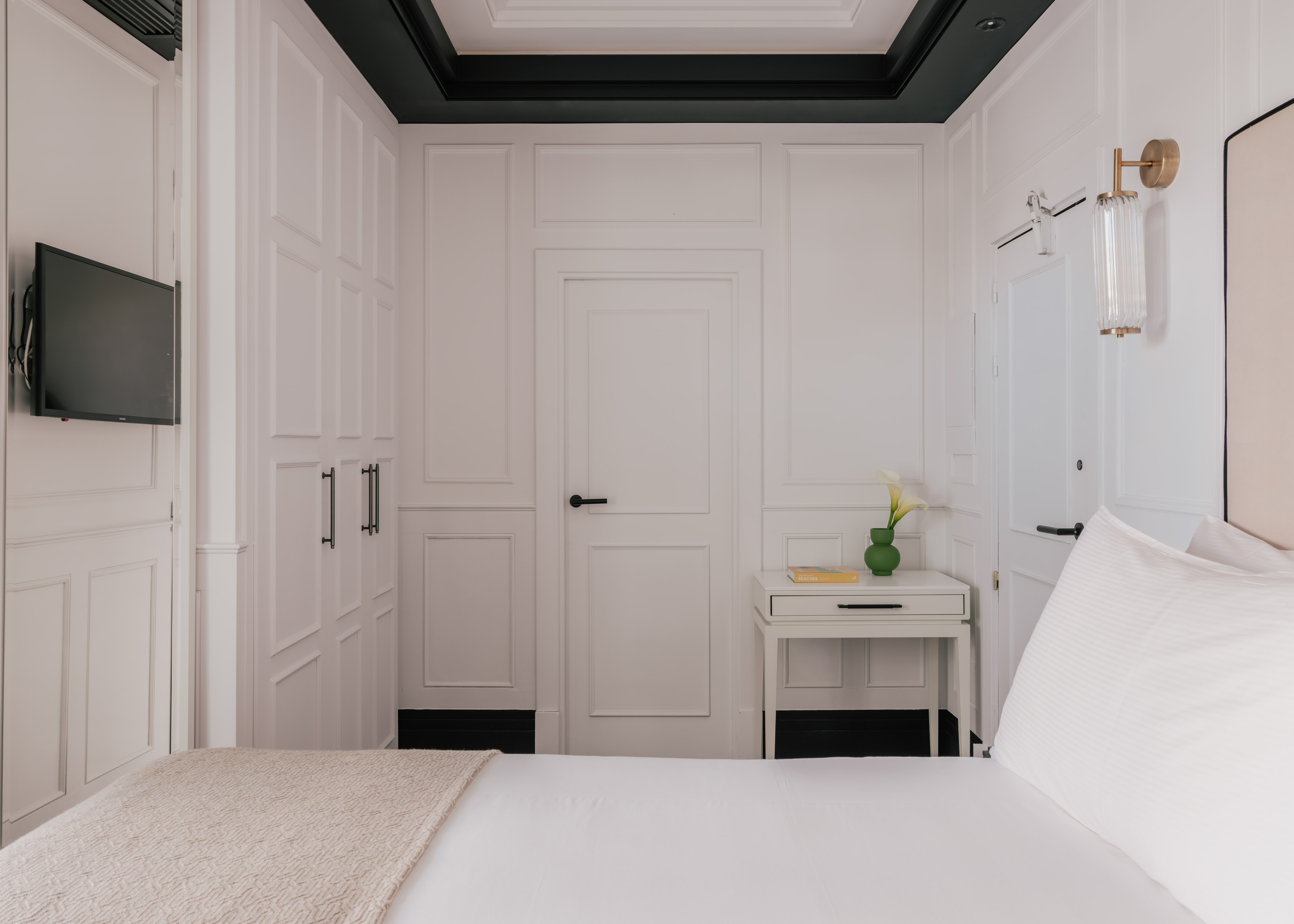Modern white bedroom with paneled walls, bed, TV, nightstand, and wall sconce.