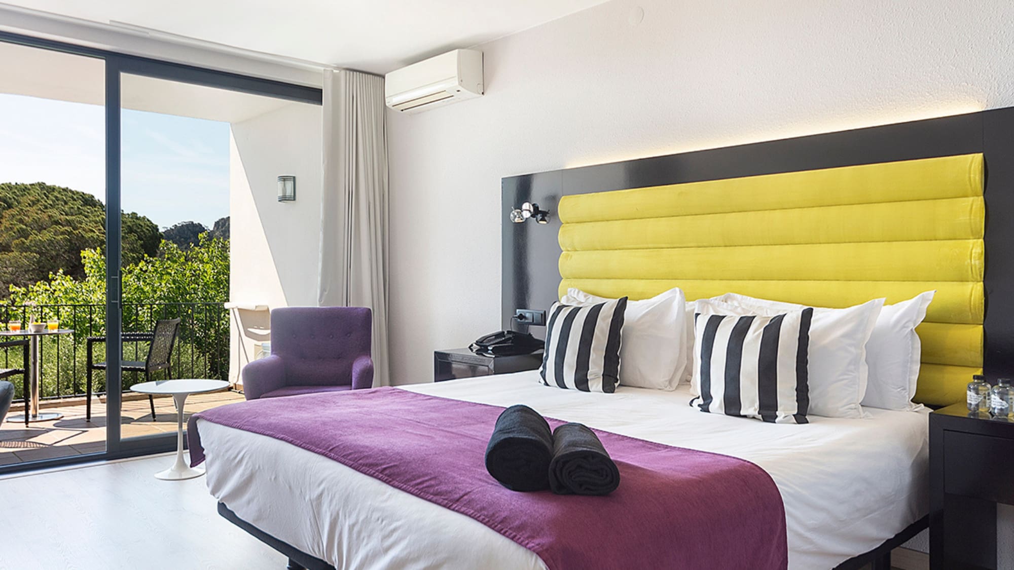 Hotel bedroom with yellow headboard, purple bed, and balcony forest view.