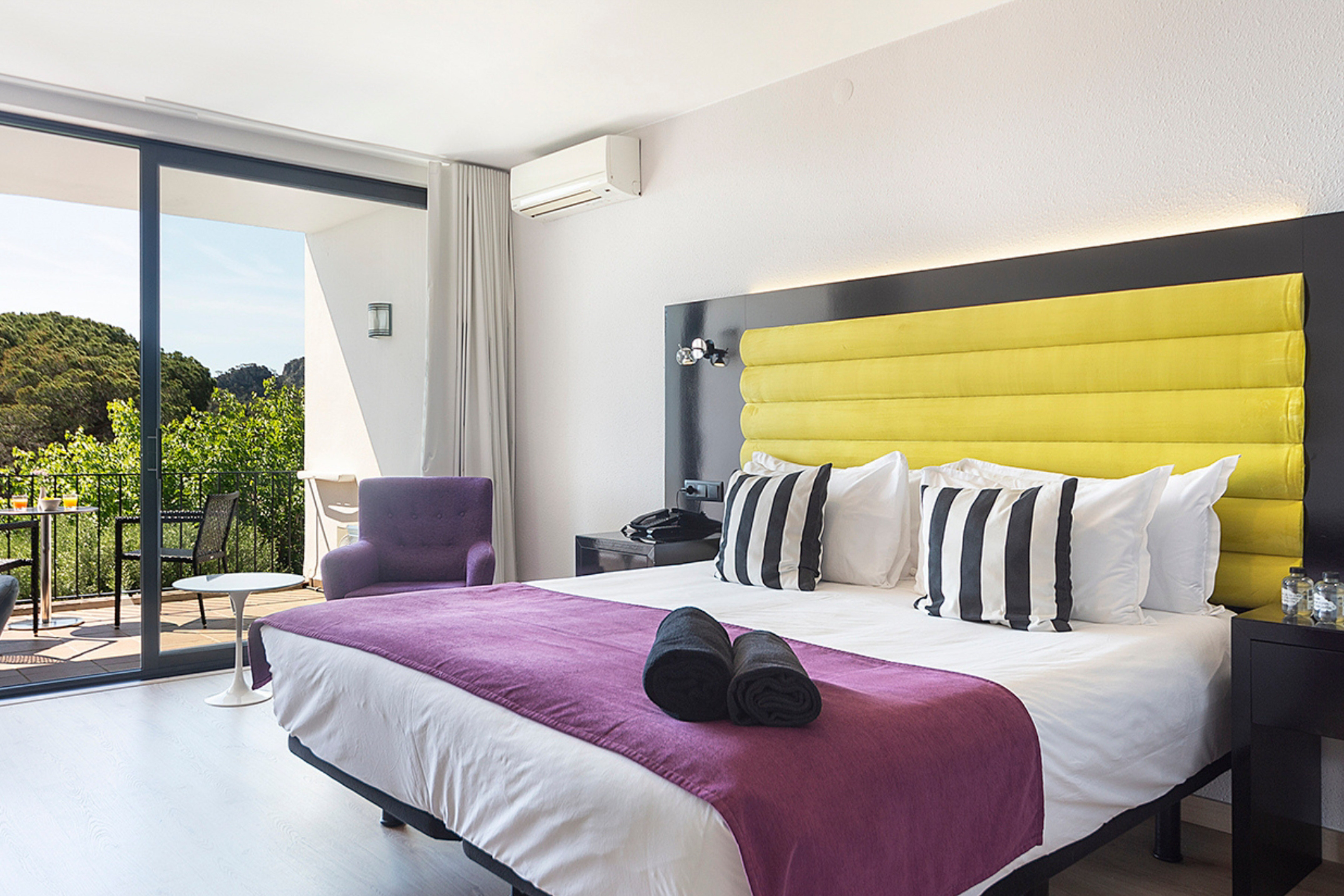 Hotel bedroom with yellow headboard, purple bed, and balcony forest view.