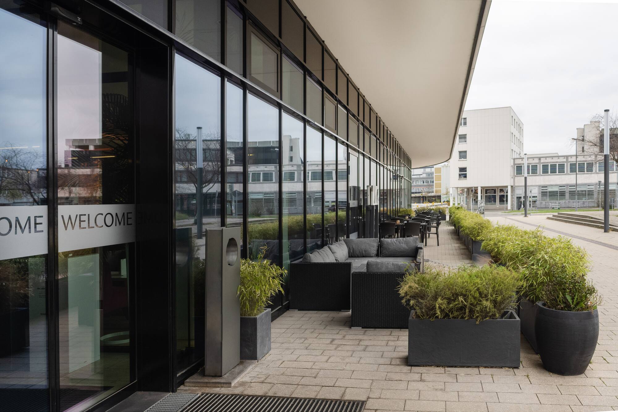 Outdoor cafe terrace with modern seating, planters, "WELCOME" on glass building.