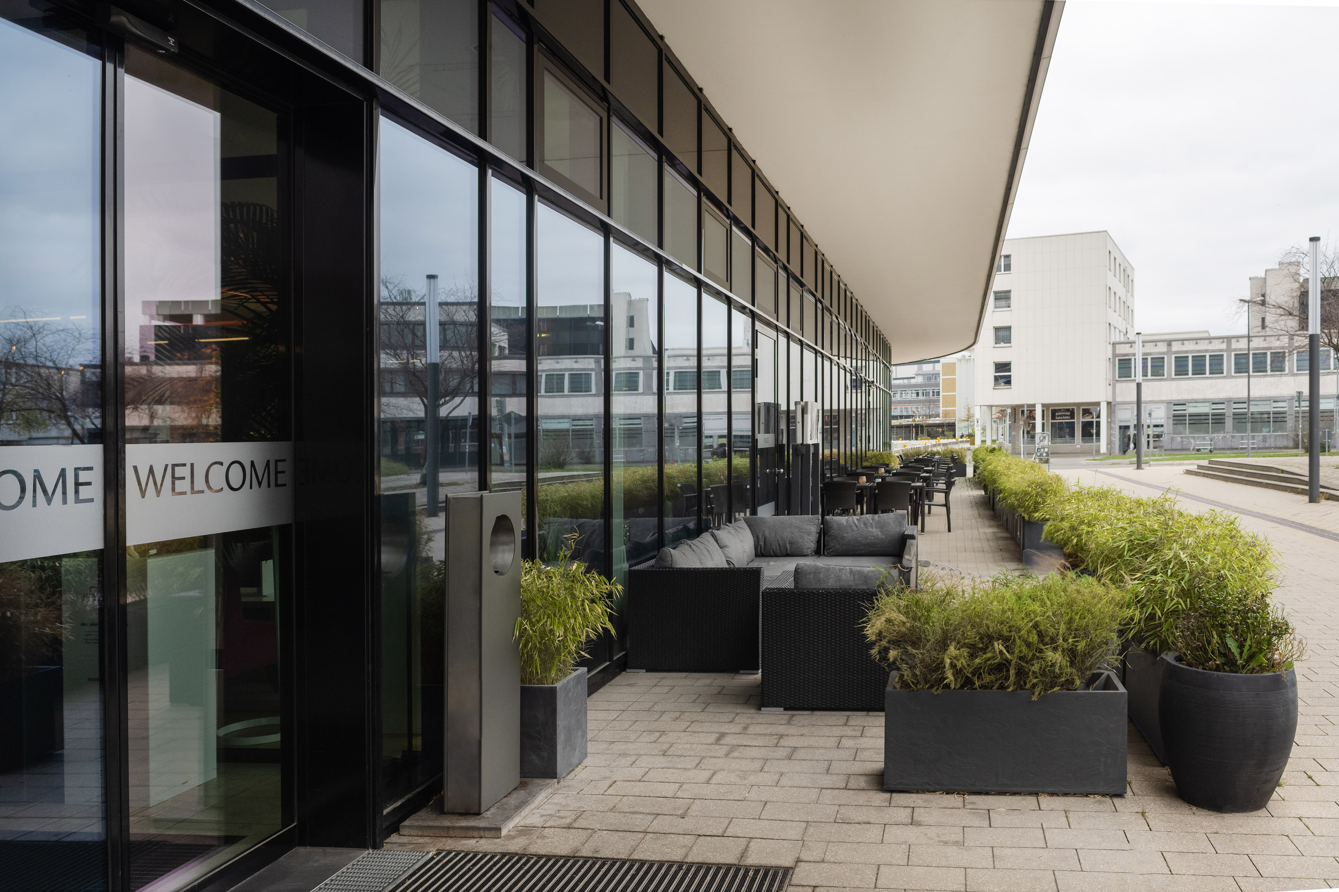 Outdoor cafe terrace with modern seating, planters, "WELCOME" on glass building.