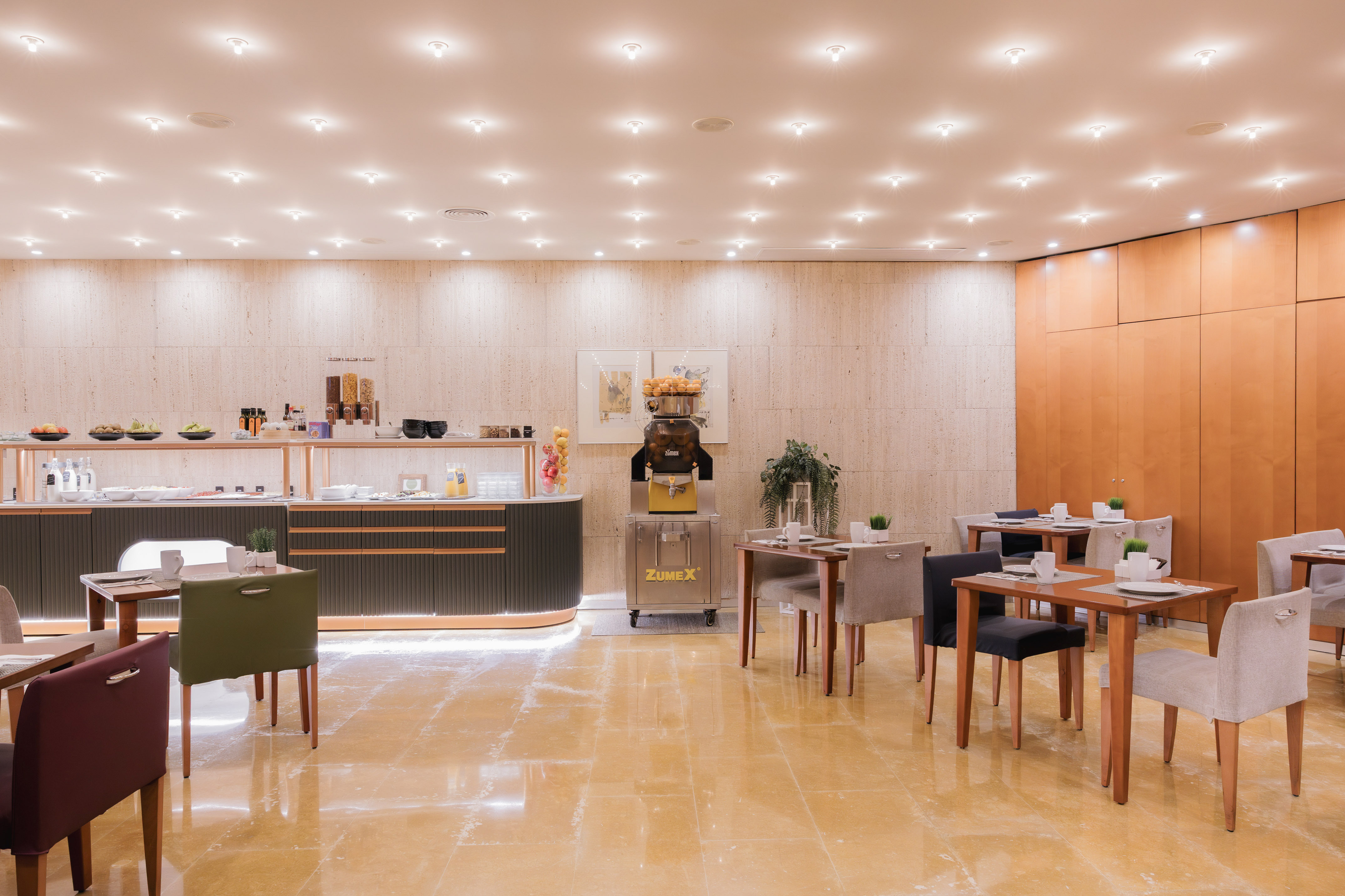 Modern hotel breakfast buffet with fresh orange juice machine and dining tables.
