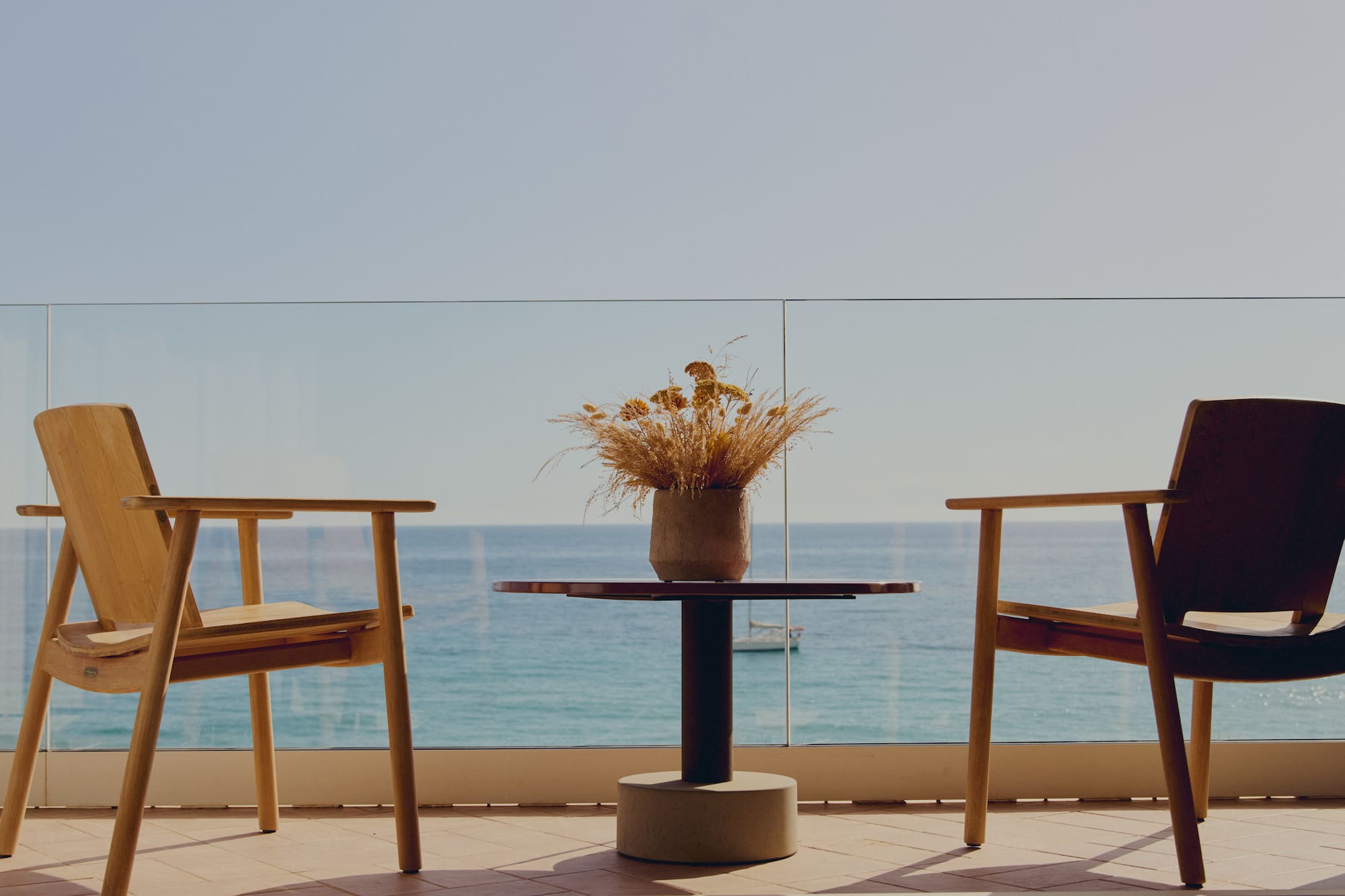 Balcón con sillas de madera, mesa y flores secas, frente al mar.