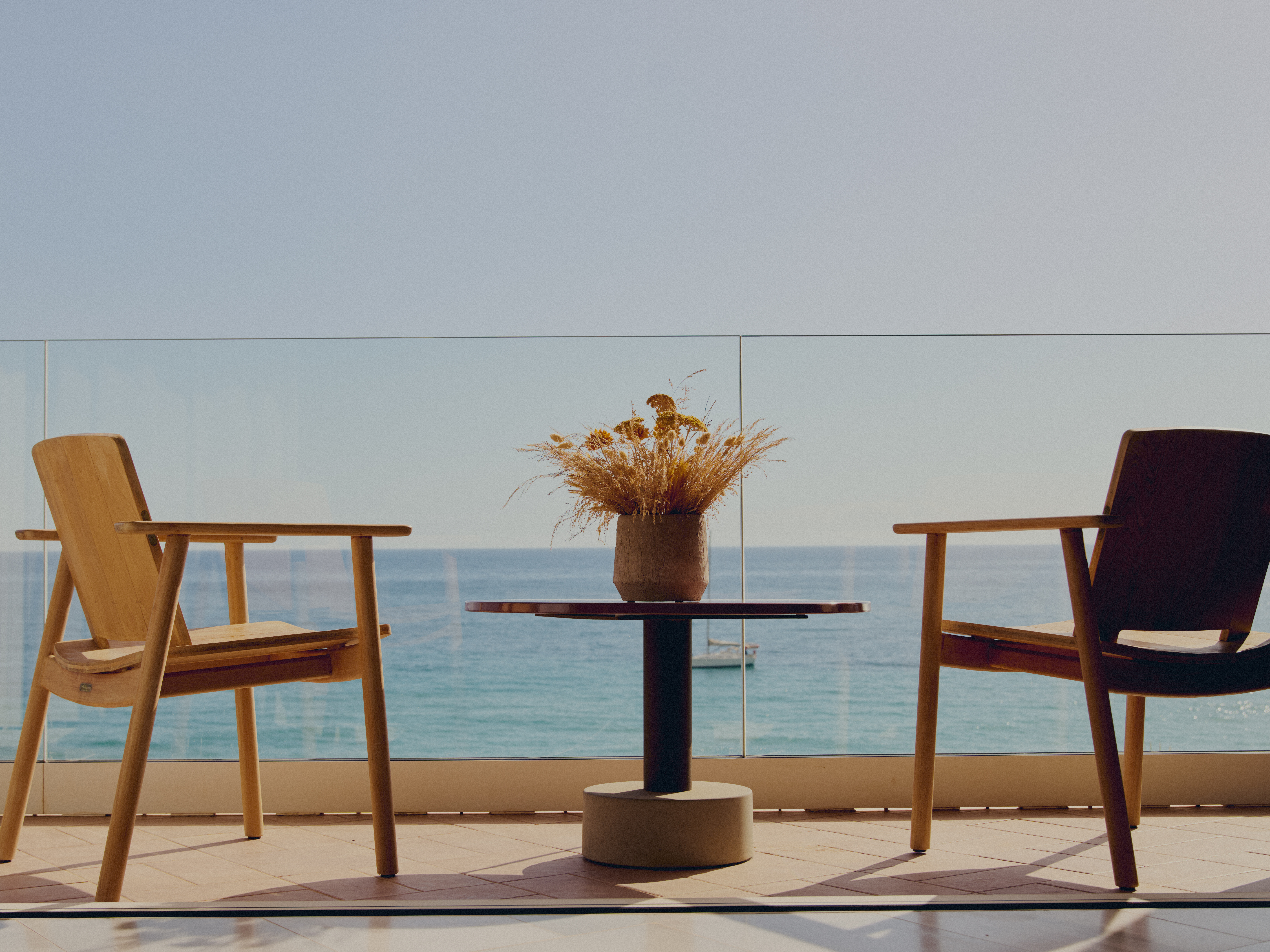 Balcón con sillas de madera, mesa y flores secas, frente al mar.