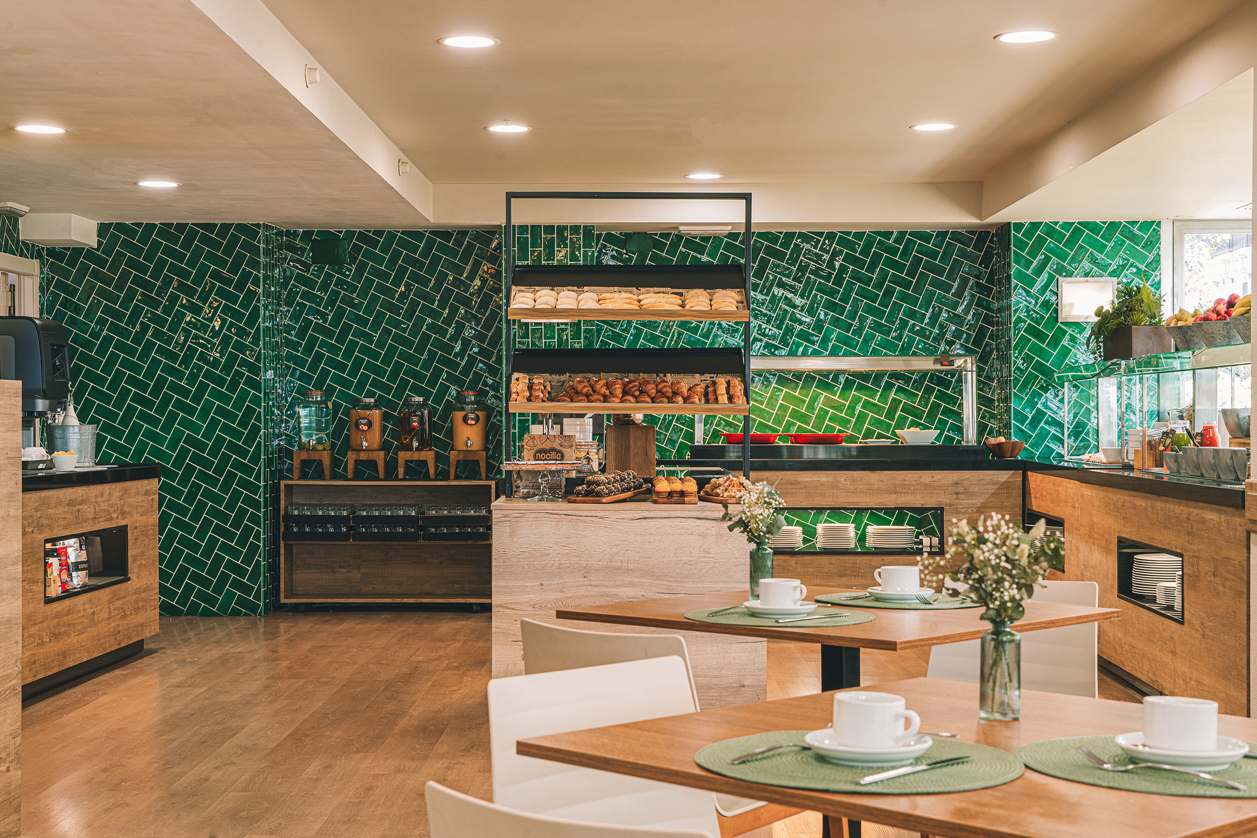 Bright breakfast buffet with green tiles, fresh pastries, juice dispensers, and dining tables.