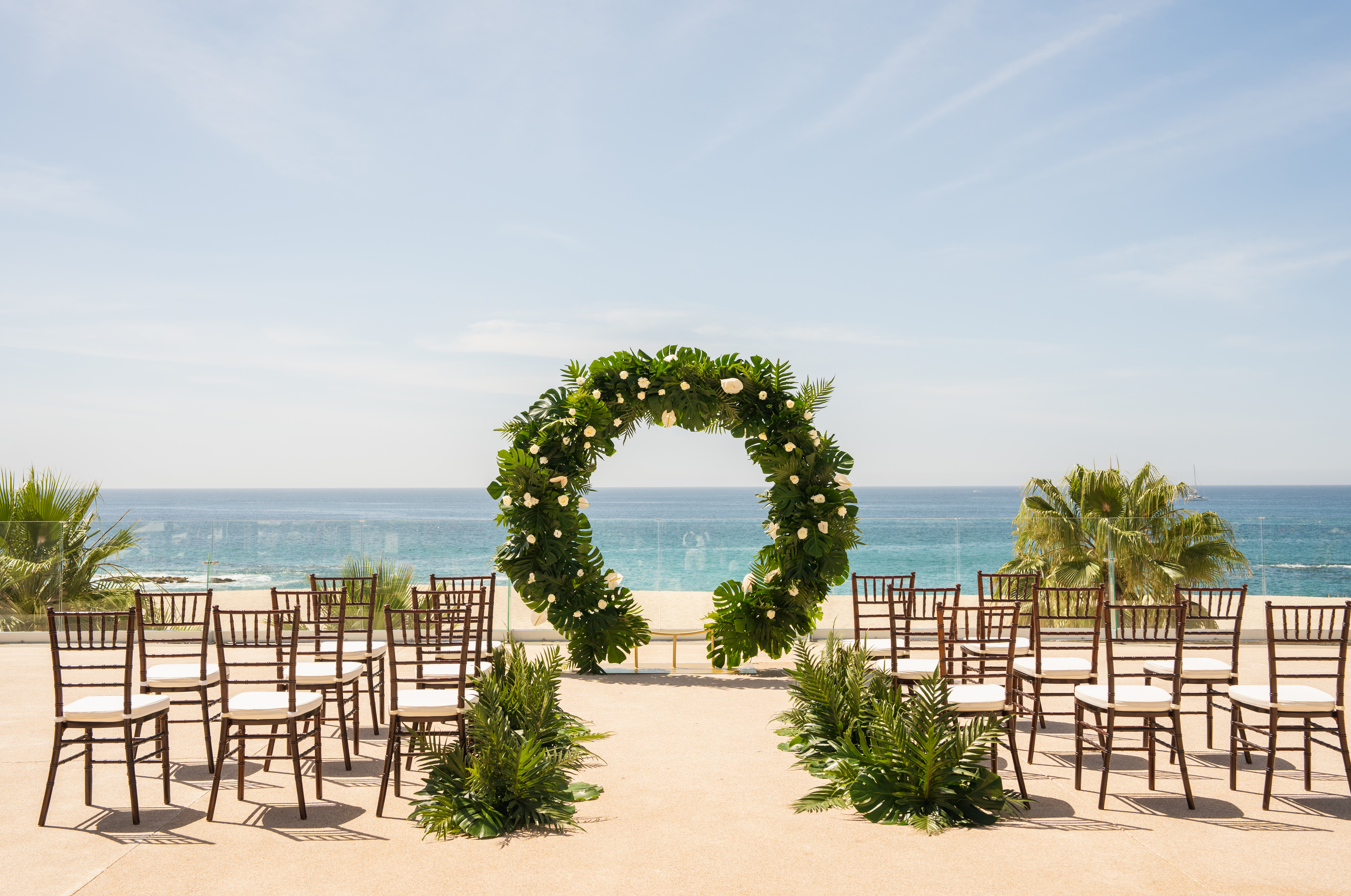 Outdoor wedding ceremony with tropical floral arch, chairs, and ocean backdrop.