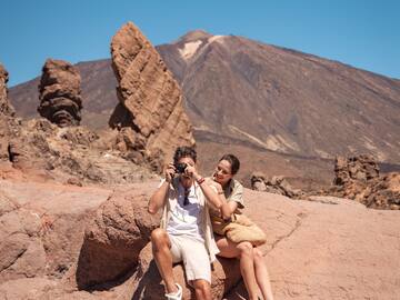 a man and woman sitting on a rock taking a picture of a mountain