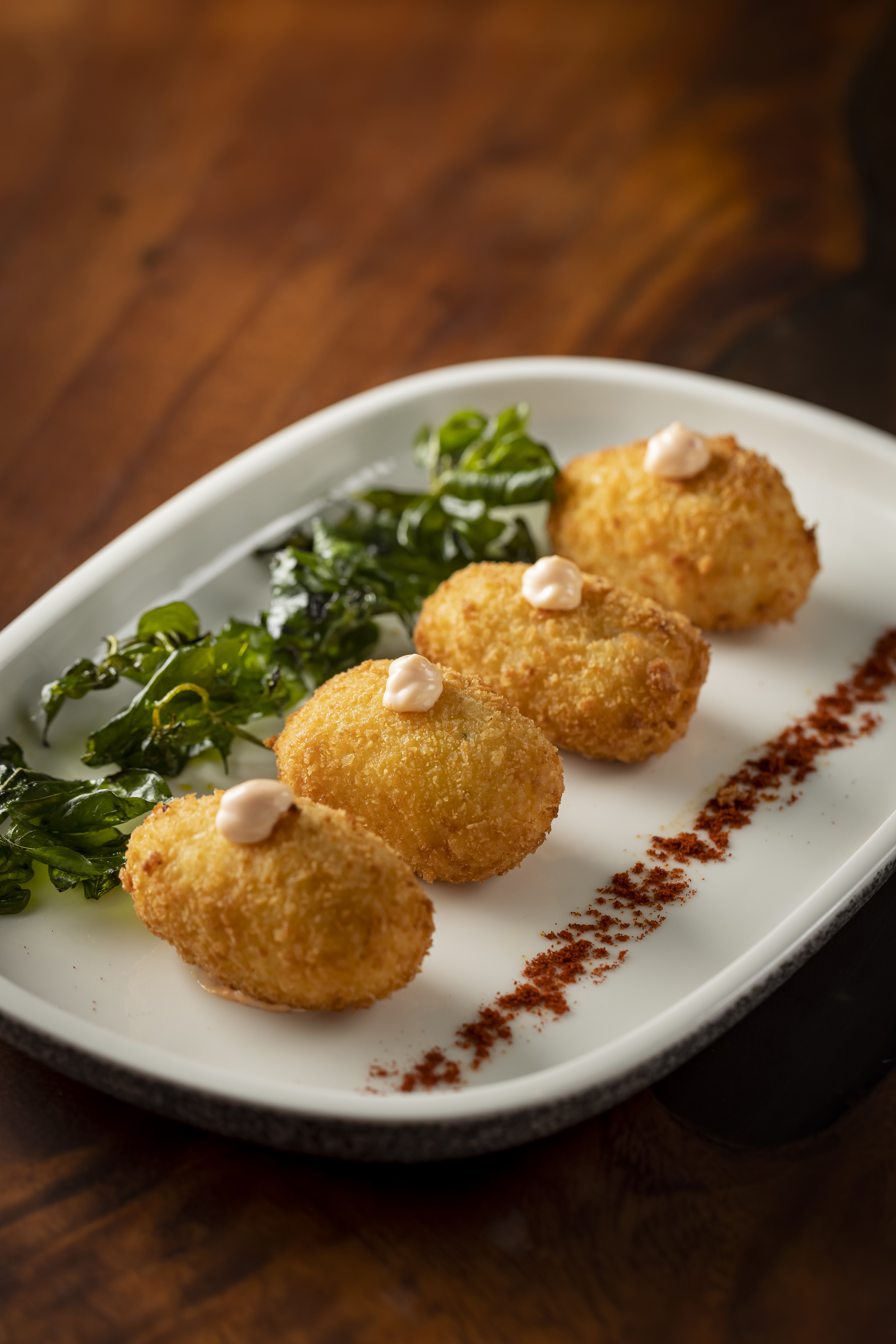 Four golden fried croquettes with sauce, greens, and paprika on white plate.
