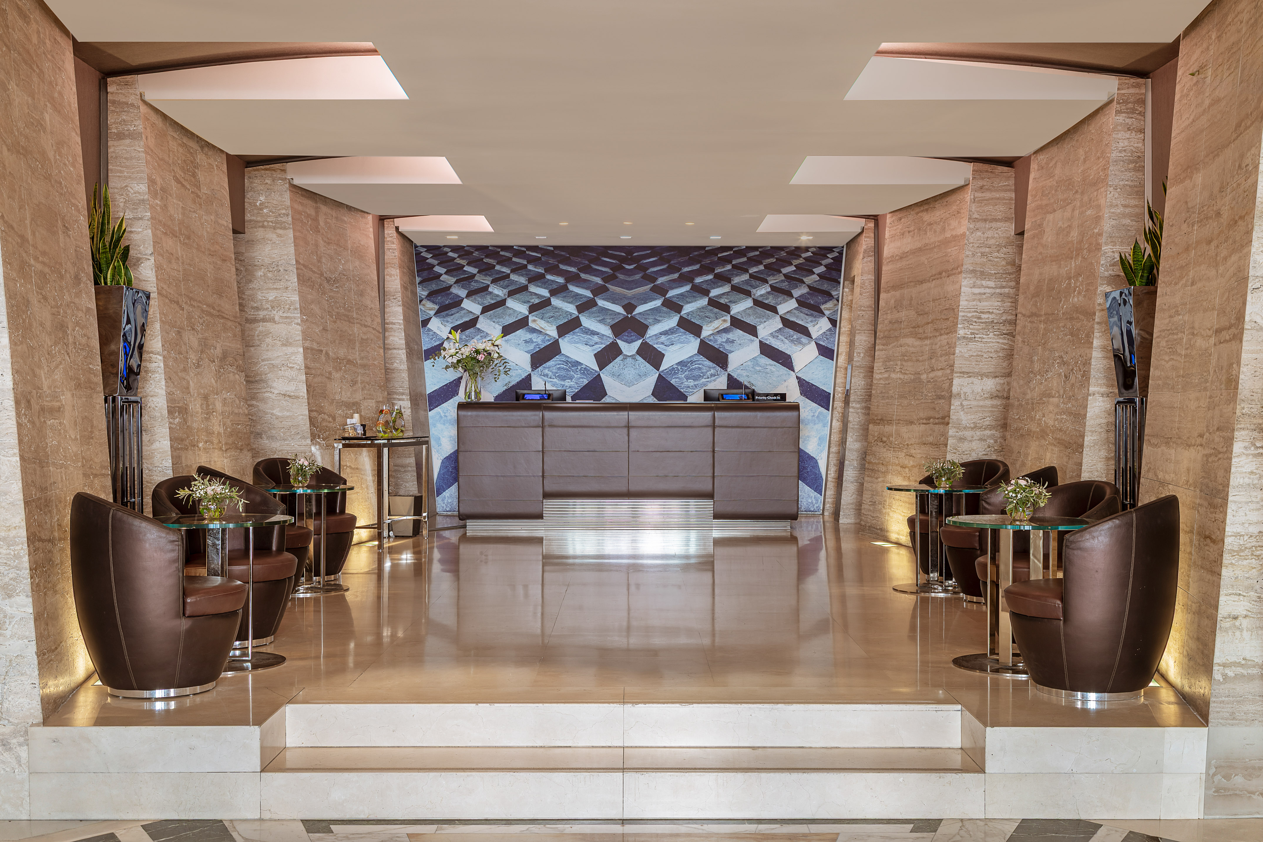 Modern hotel lobby with reception desk, blue geometric wall, and brown lounge chairs.